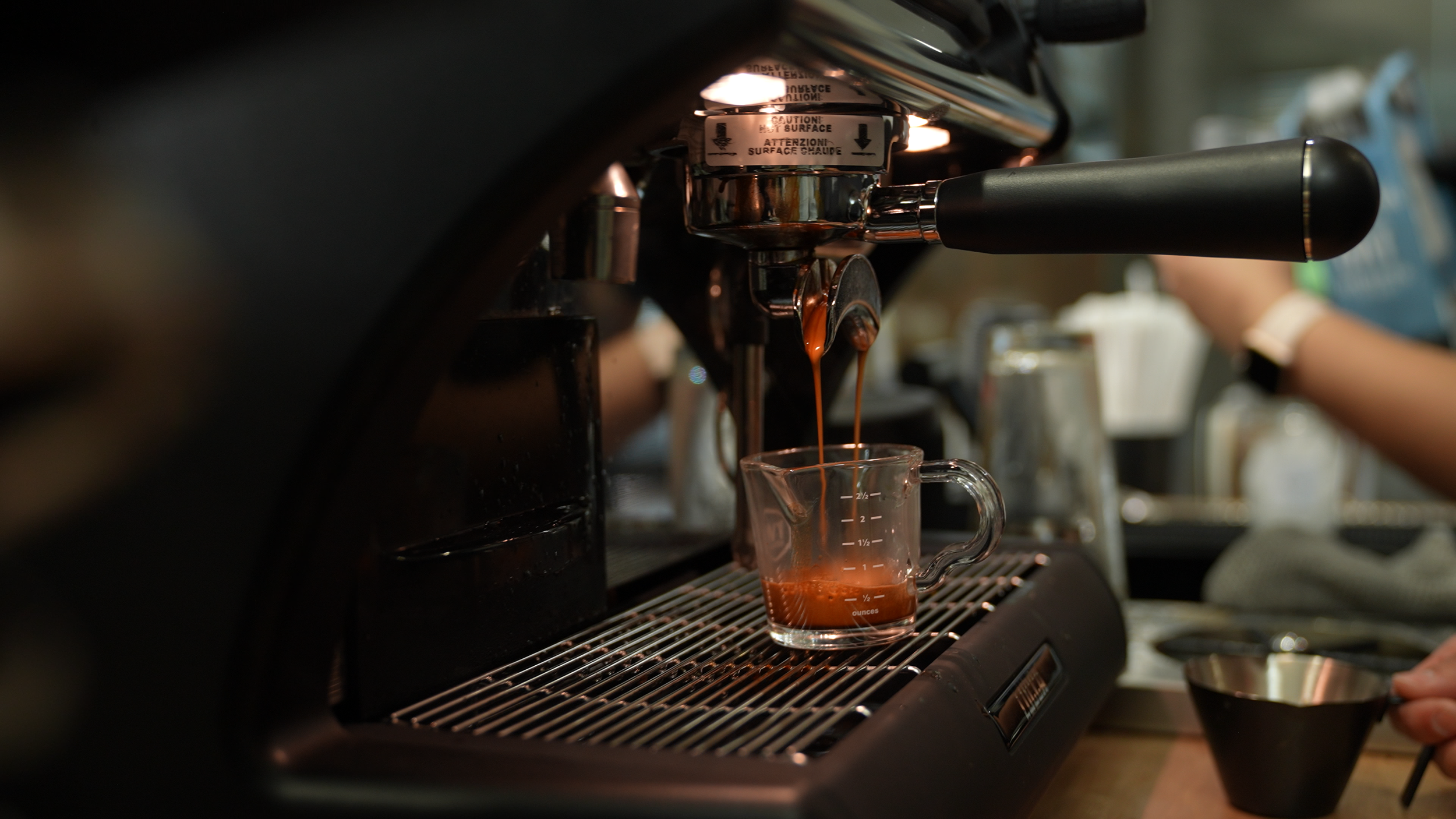 Close-up of a coffee machine pouring espresso into a glass measuring cup on a drip tray in a coffee shop setting.