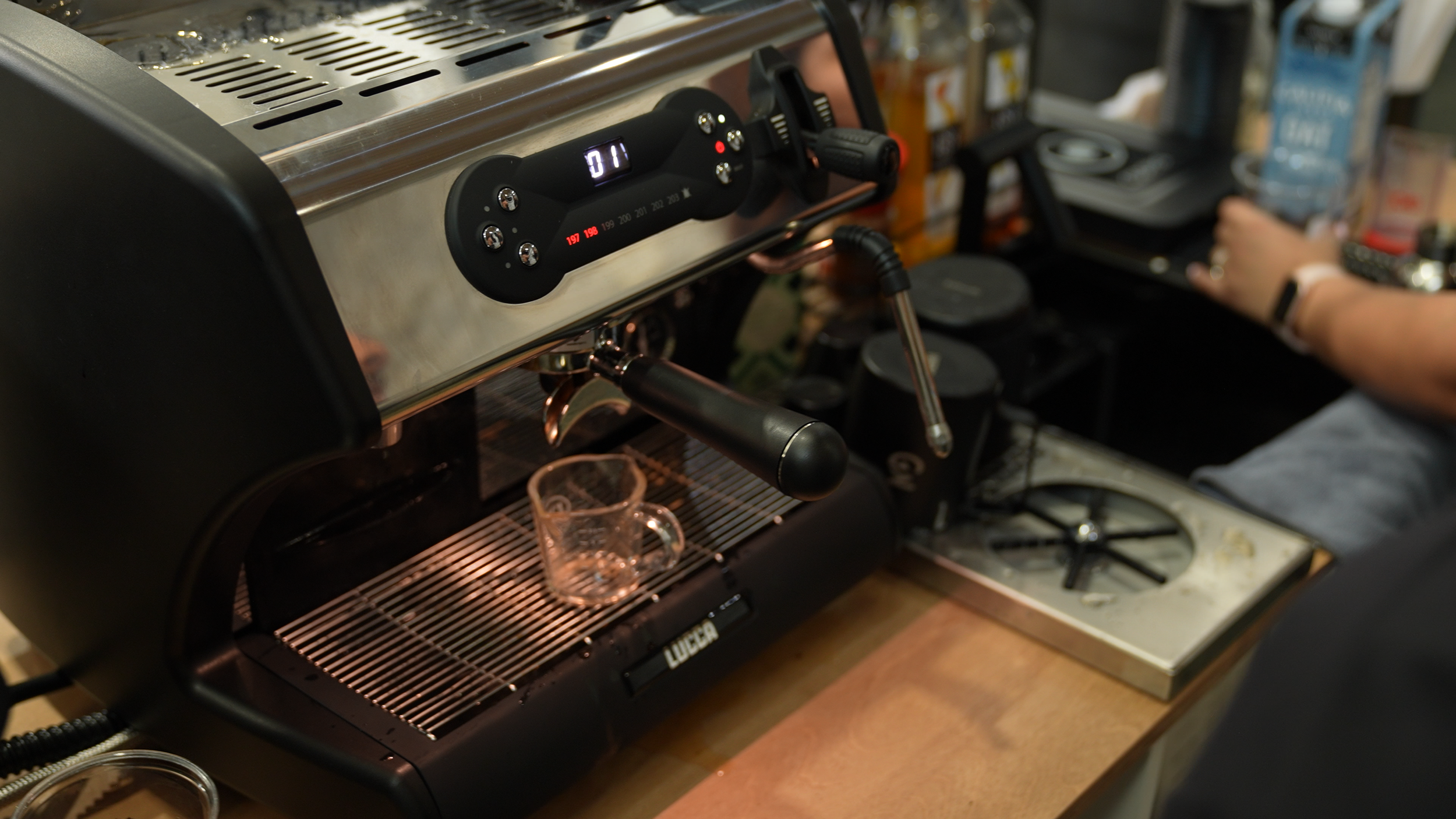 A close-up of a commercial espresso machine brewing coffee with a glass mug placed underneath, and a barista's hand adjusting something in the background.
