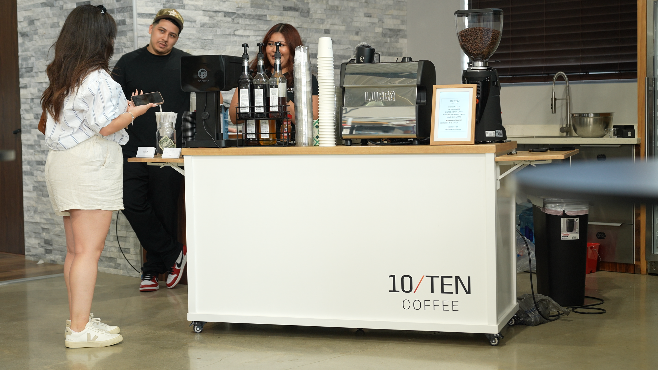 A customer orders at a coffee stand inside a modern cafe, with two baristas behind the counter preparing coffee.