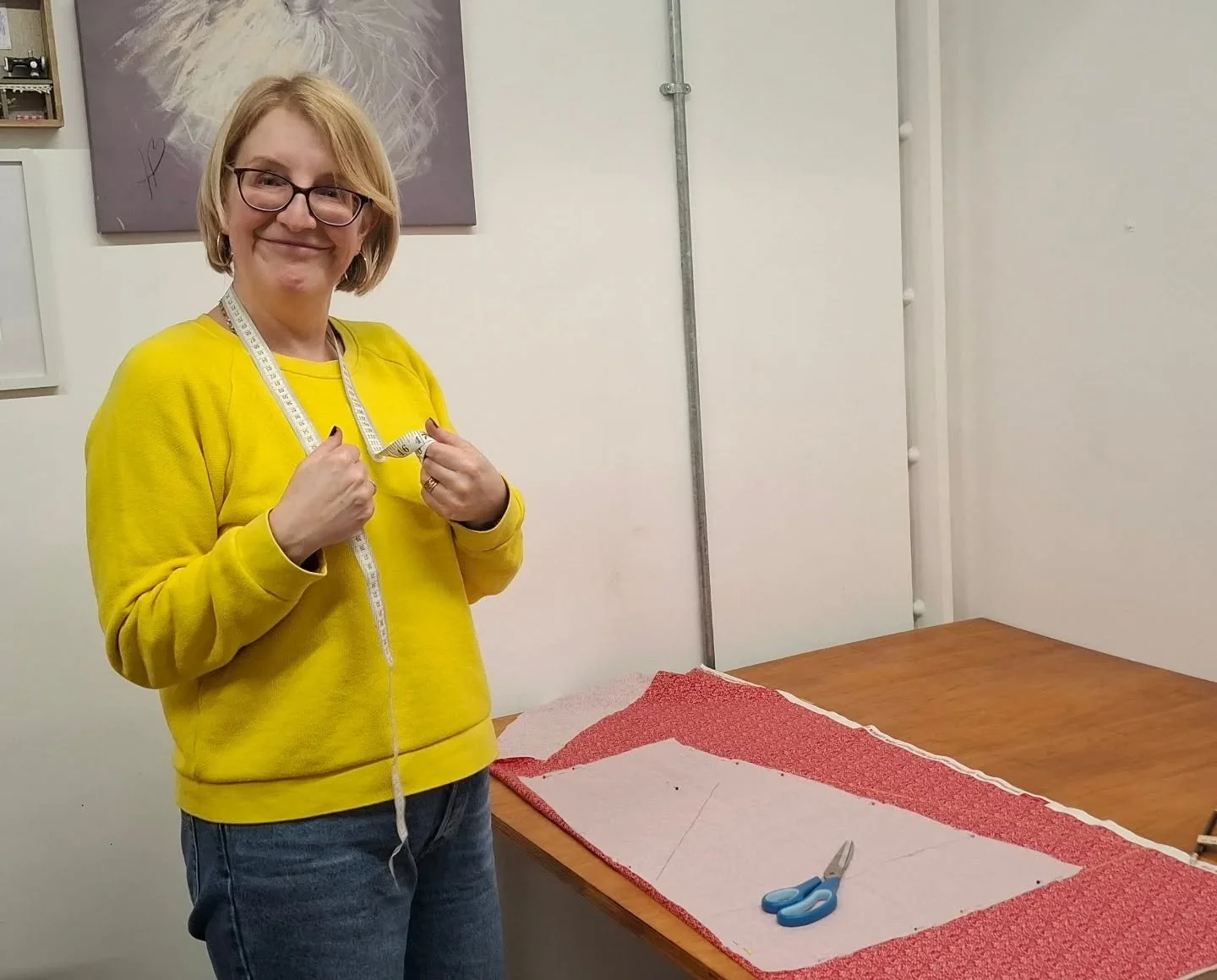 A woman in a yellow sweatshirt holding a measuring tape around her neck, standing next to a wooden table covered with fabric and a pair of scissors.