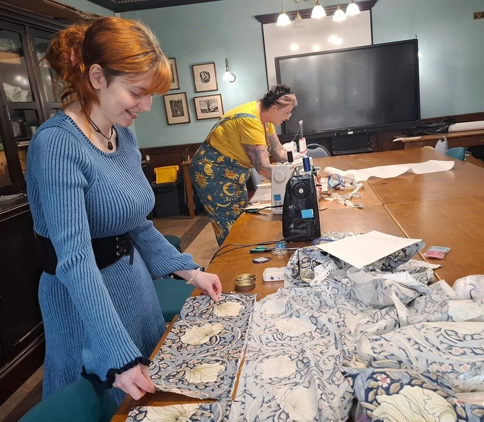 Two women working on sewing projects in a workshop room, with fabric and sewing tools on the table.