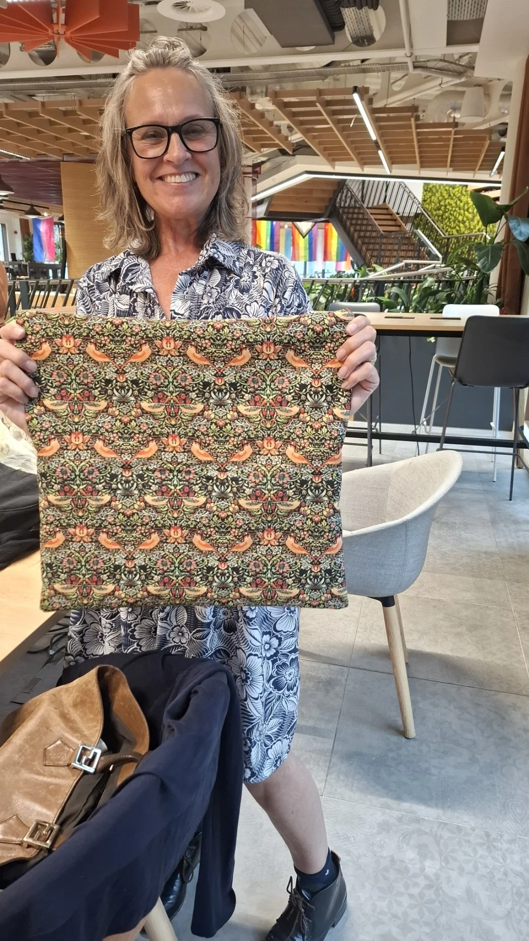 A woman with glasses and shoulder-length hair smiling and holding up a decorated fabric in a modern, colorful indoor space.