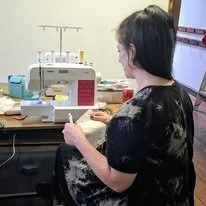 A woman sitting at a table using a sewing machine. The table has sewing supplies and fabric. The woman is facing away from the camera.