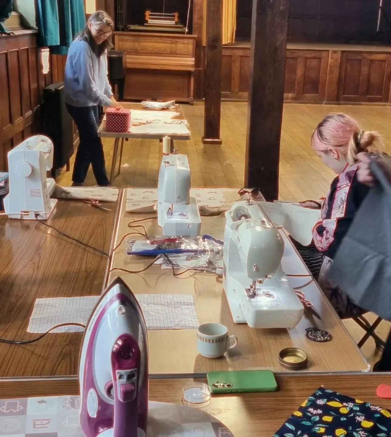 Two women working with sewing machines on a wooden table, surrounded by sewing supplies and an iron, in a room with wooden walls and floors.