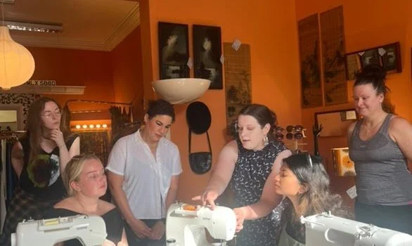 A group of six women gathered around a table, with sewing machines, in a warmly lit room with orange walls and various framed pictures and decorations.