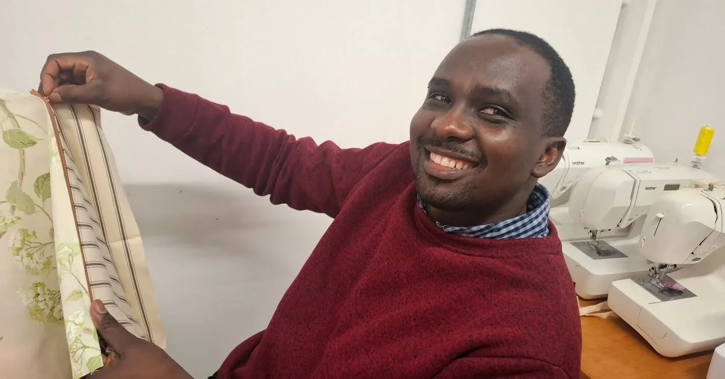 A man smiling and sewing on a white sewing machine with multiple other sewing machines in the background.