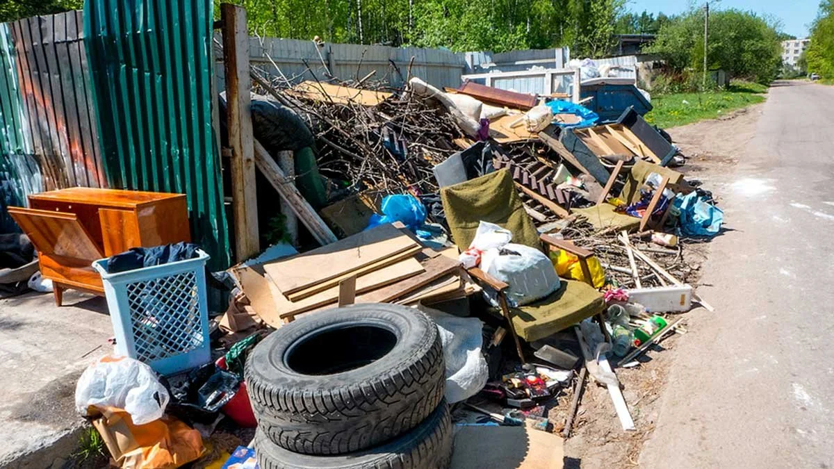 A large pile of trash and debris along a sidewalk, including tires, furniture, cardboard, plastic bags, and various household items, next to a green metal fence on a street with trees in the background.