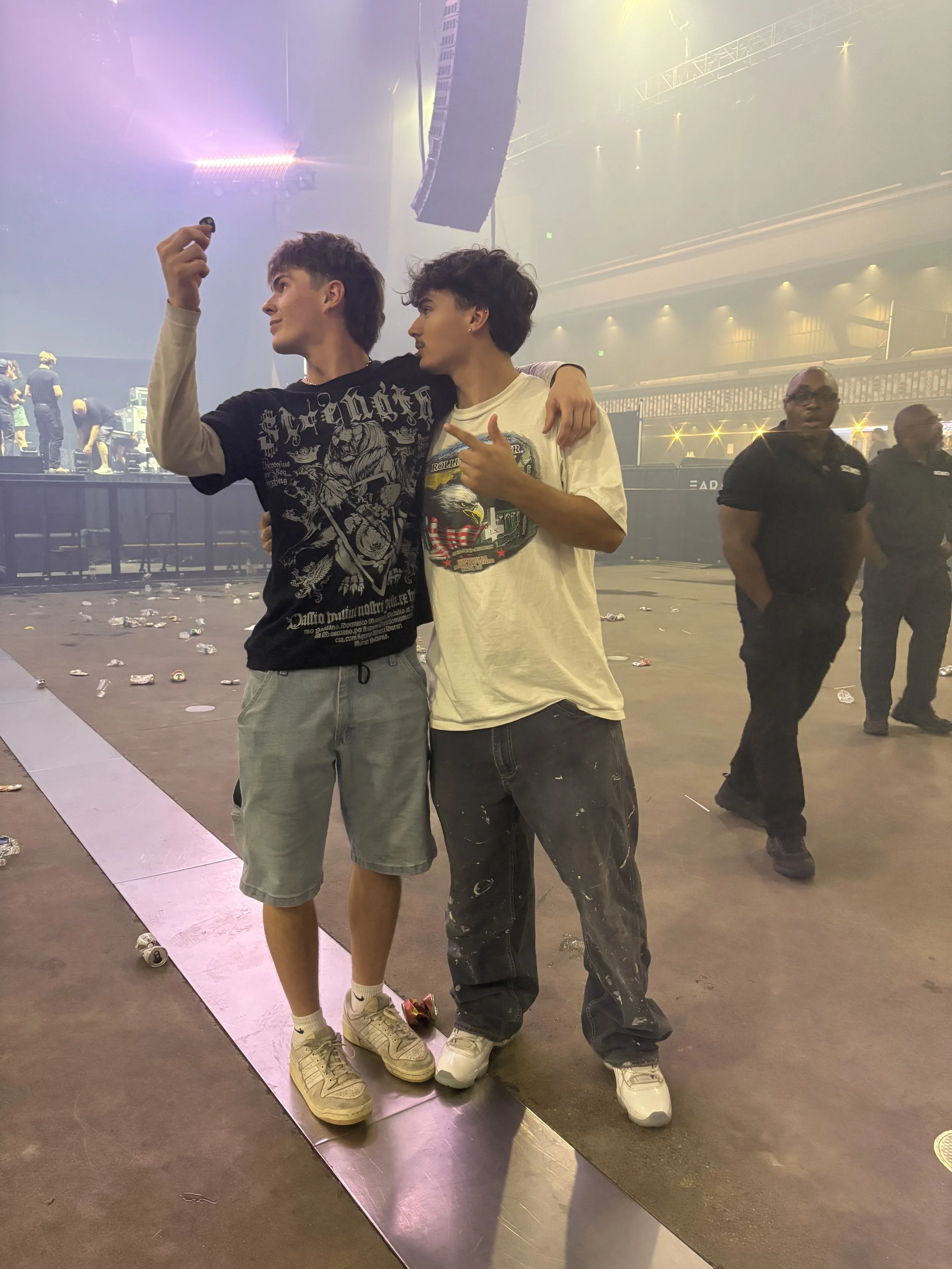 Two young men taking a selfie at a concert or music event. They are standing close, with one having his arm around the other, on a floor strewn with debris and empty cans. Stage lights and other people in the background suggest an active concert environment.