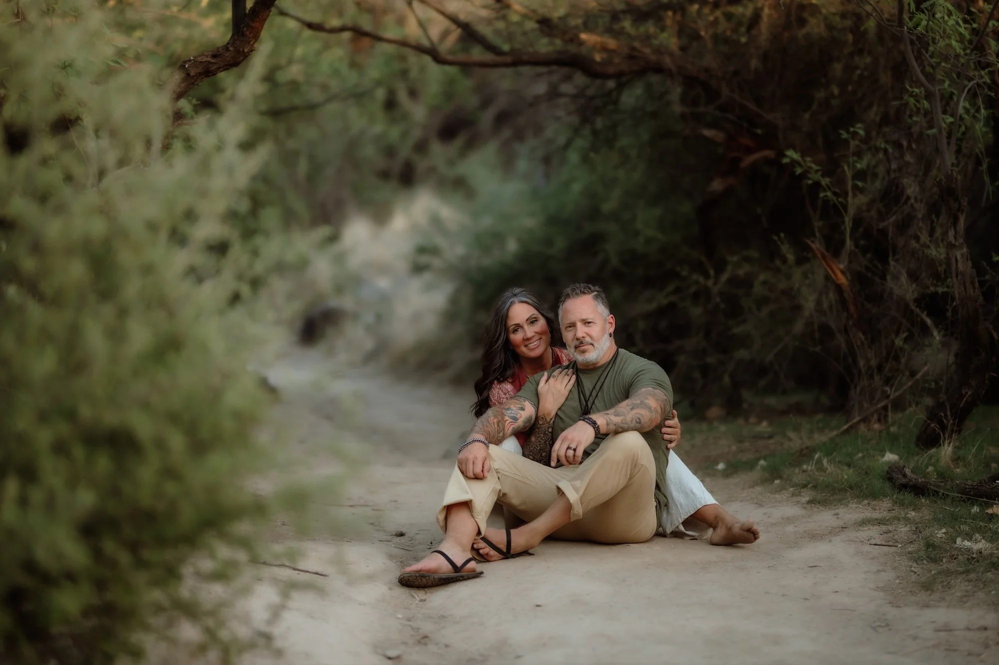 A couple sitting on a dirt path surrounded by trees and bushes.