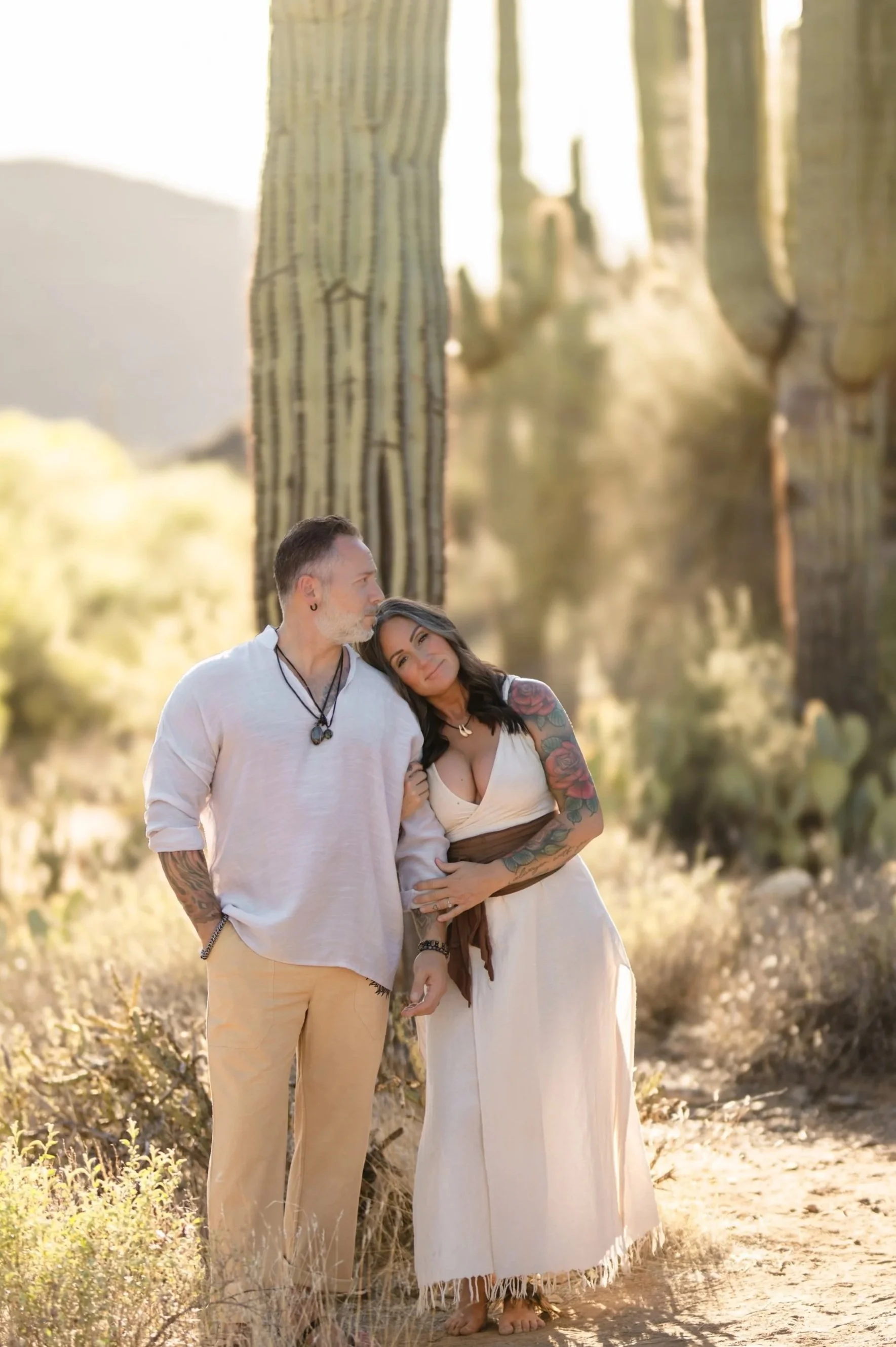 A man and a woman with tattoos standing close together in a desert landscape with large cacti, during sunset.