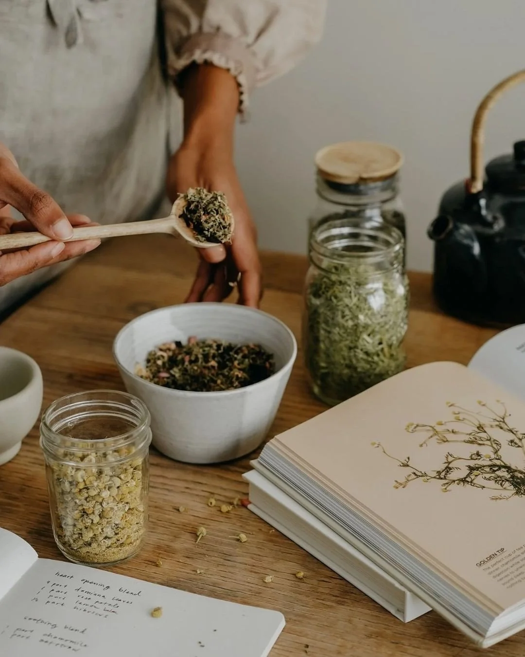 Person scooping dried herbs from a jar into a bowl on a wooden table, with additional jars, a teapot, a cup, a notebook, and herbs on the table.