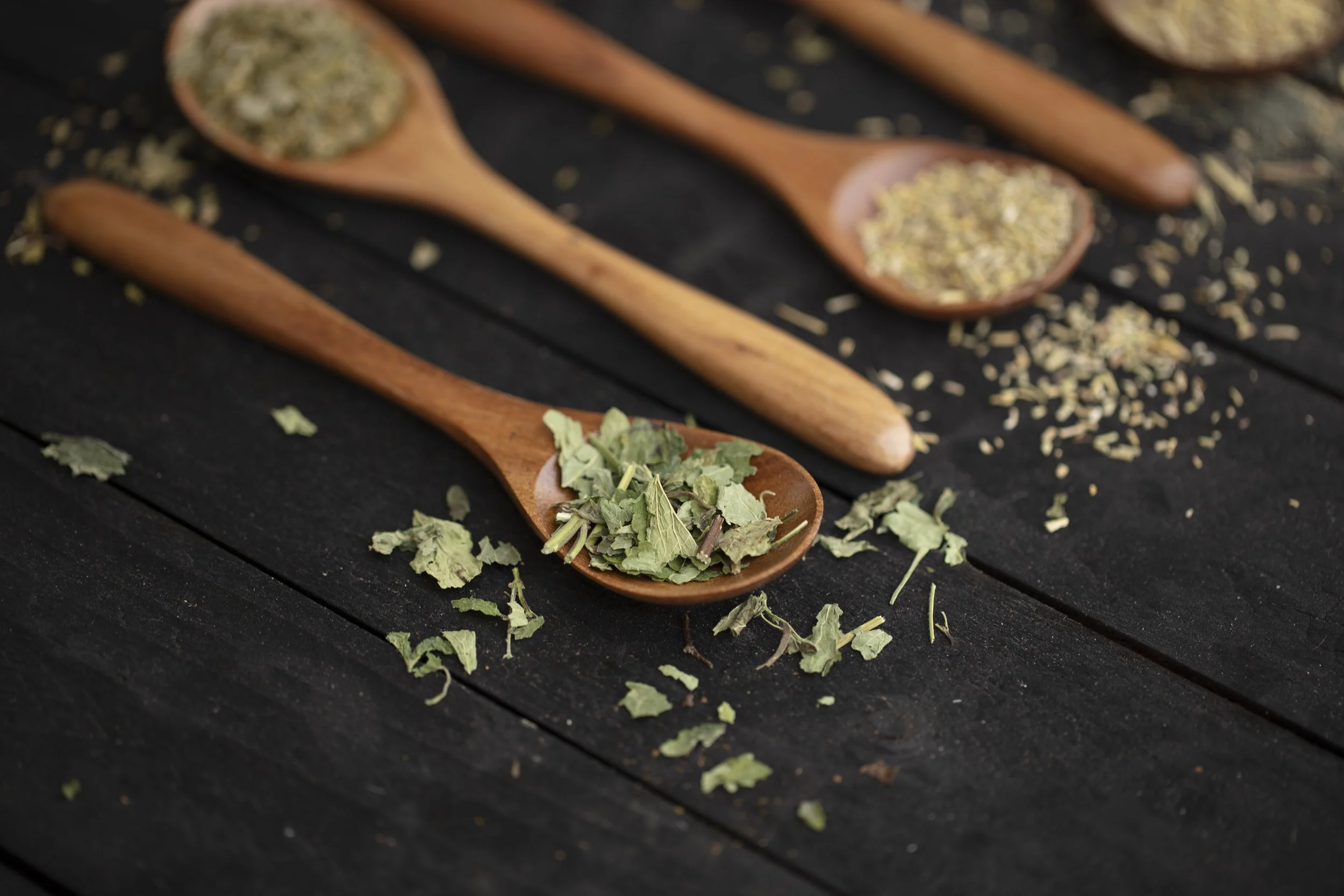 Three wooden spoons on a dark wooden surface, each filled with different herbs or spices, with some herbs scattered around.