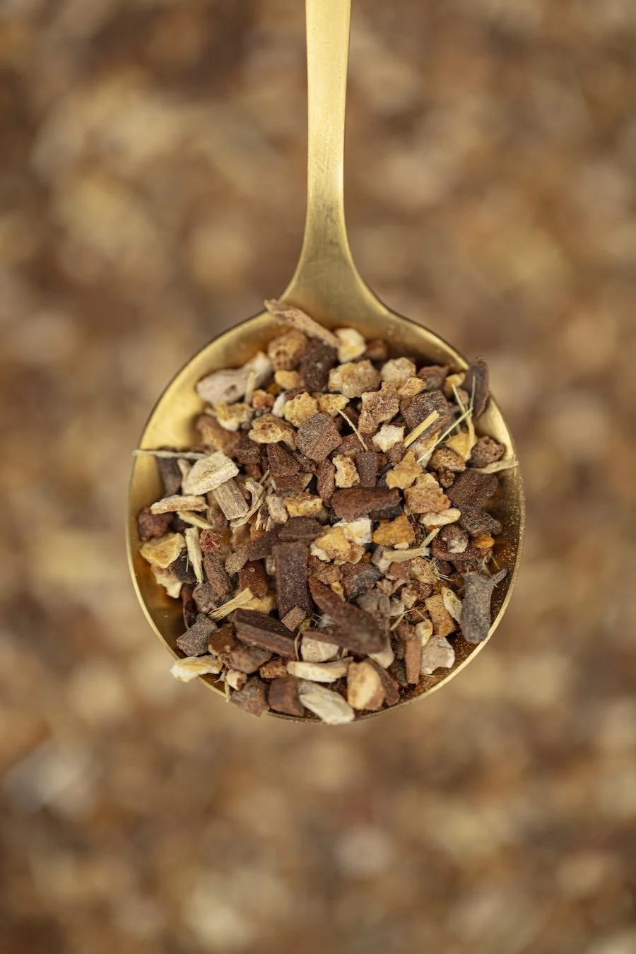 Close-up of a gold spoon filled with chopped dried herbs and spices, against a blurred background of similar herbs and spices.