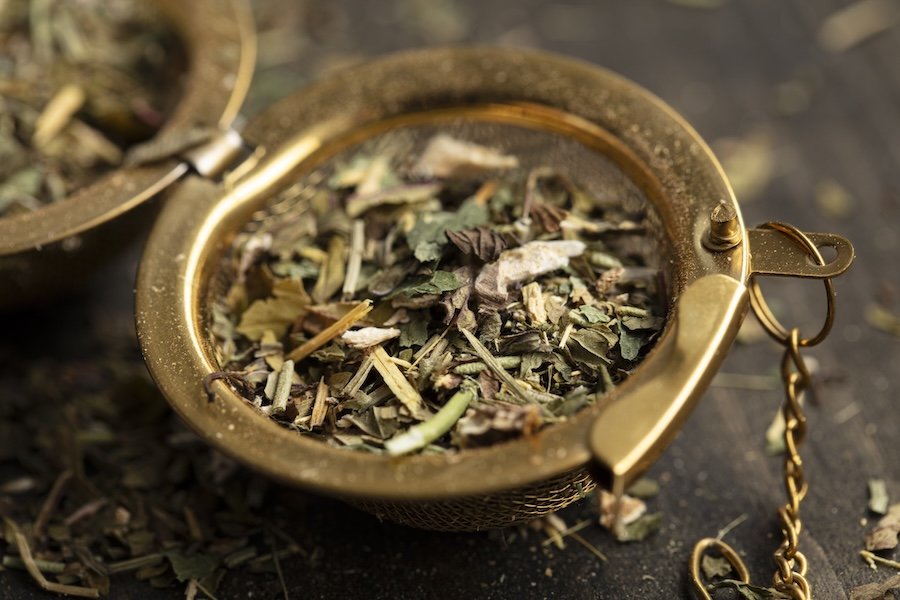 Close-up of a gold-colored metal container filled with dried herbal tea leaves, with some spilled on the dark surface below.