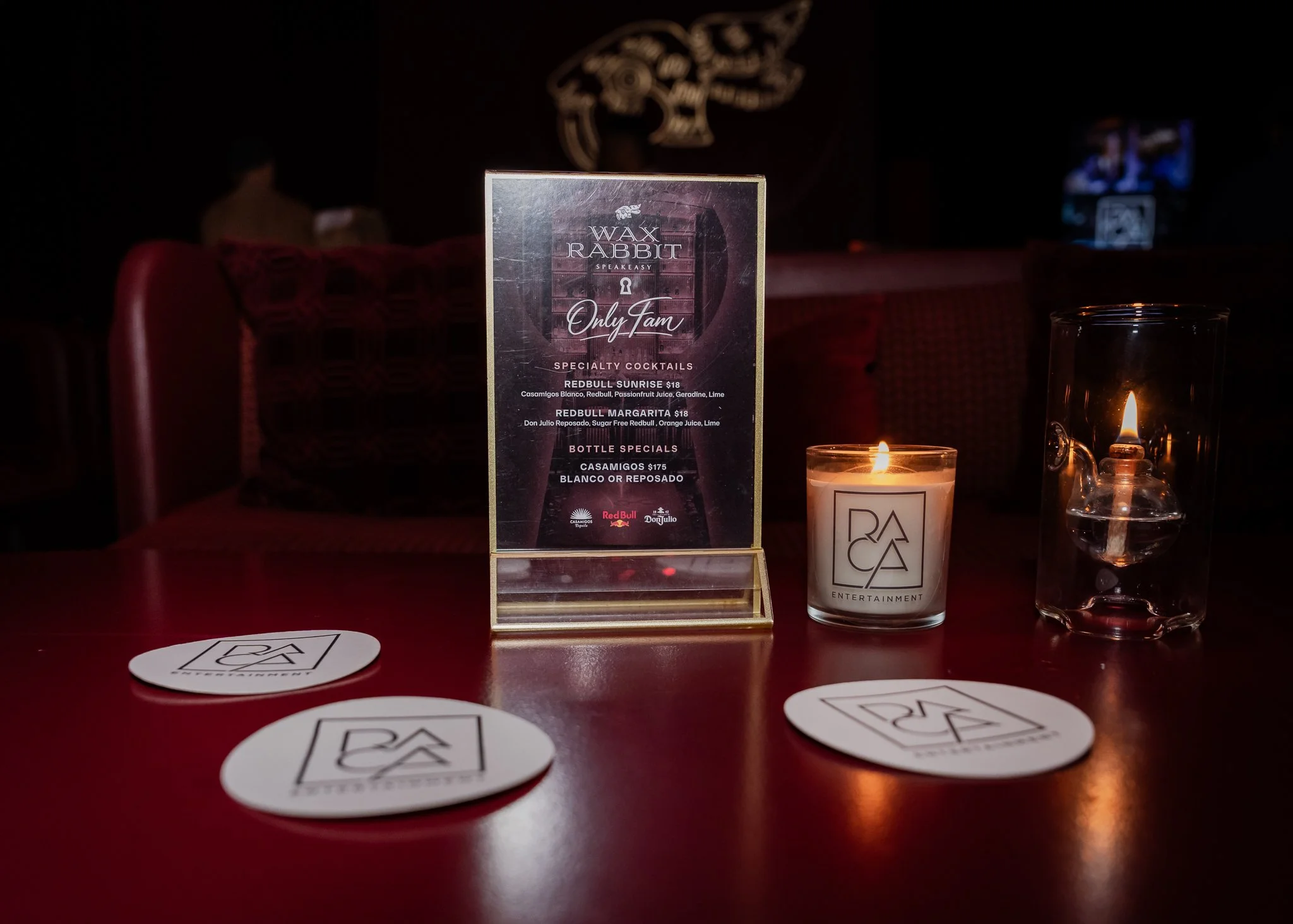 A table with a menu card for 'Wax Rabbit Speakeasy' listing specialty cocktails and bottle specials, a lit candle, and several round stickers with a logo on a red surface in a dimly lit room.