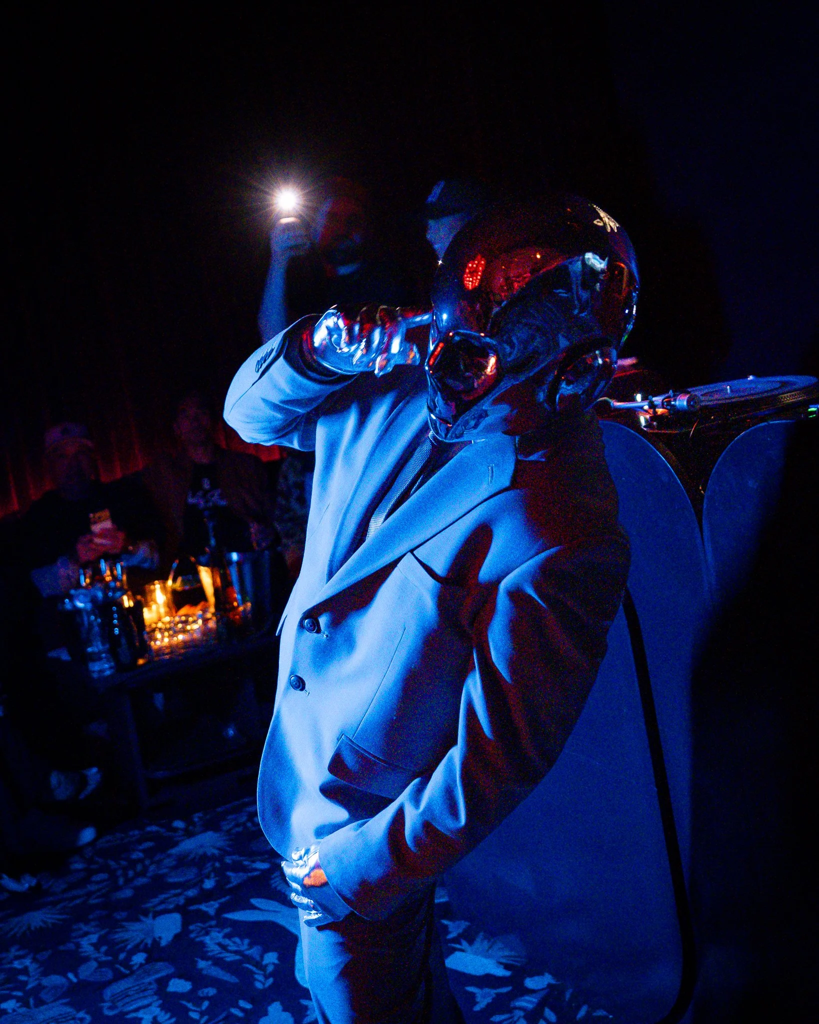 A person in a black helmet and a white jacket with red accents is sitting at a table in a dark room, possibly at night, illuminated by a bright light source behind them. The background includes a floral tablecloth and bottles of drinks on the table.