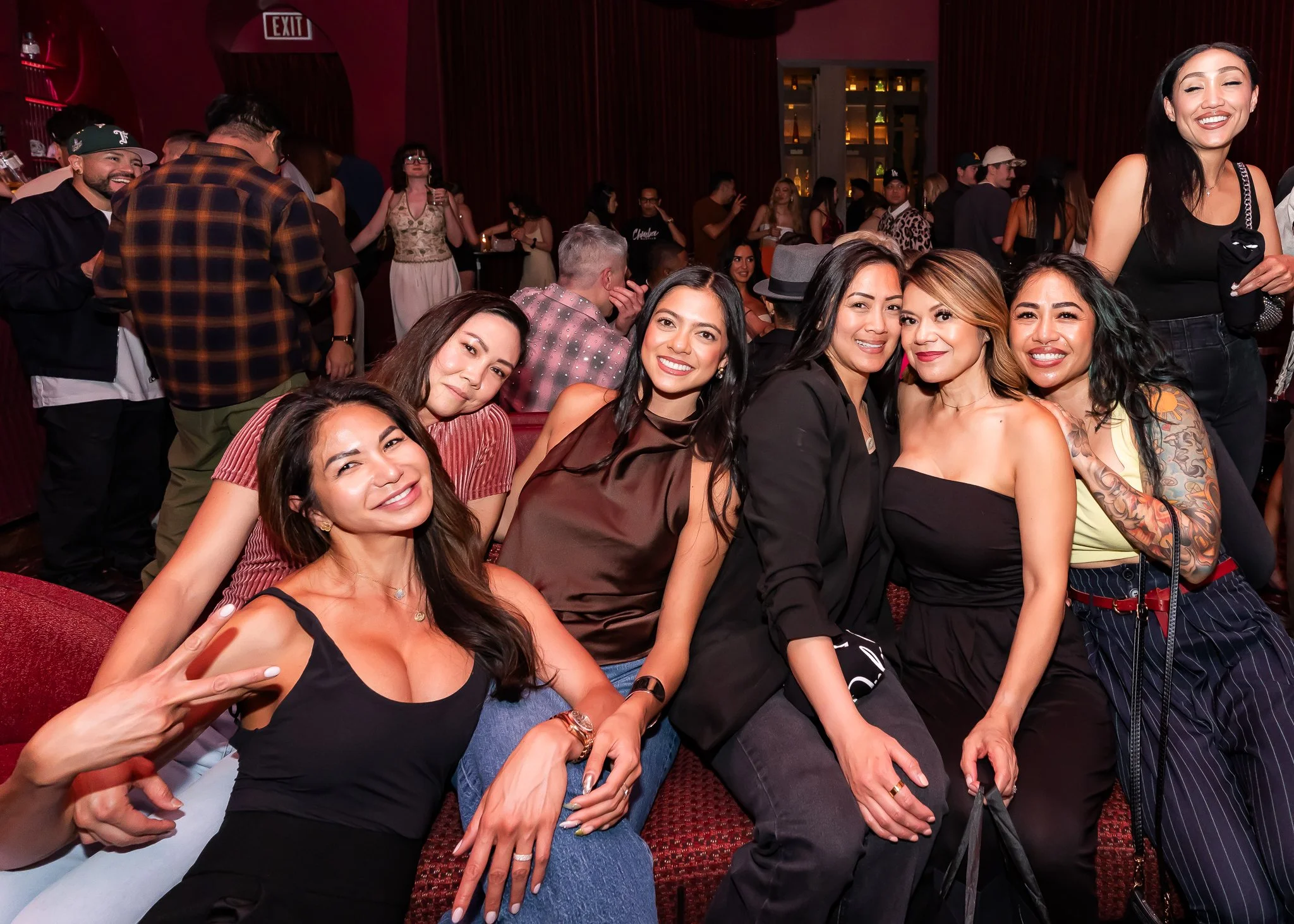 Group of women and men enjoying themselves at a party or social event inside a dimly lit venue with red walls and curtains, smiling and posing for the camera.