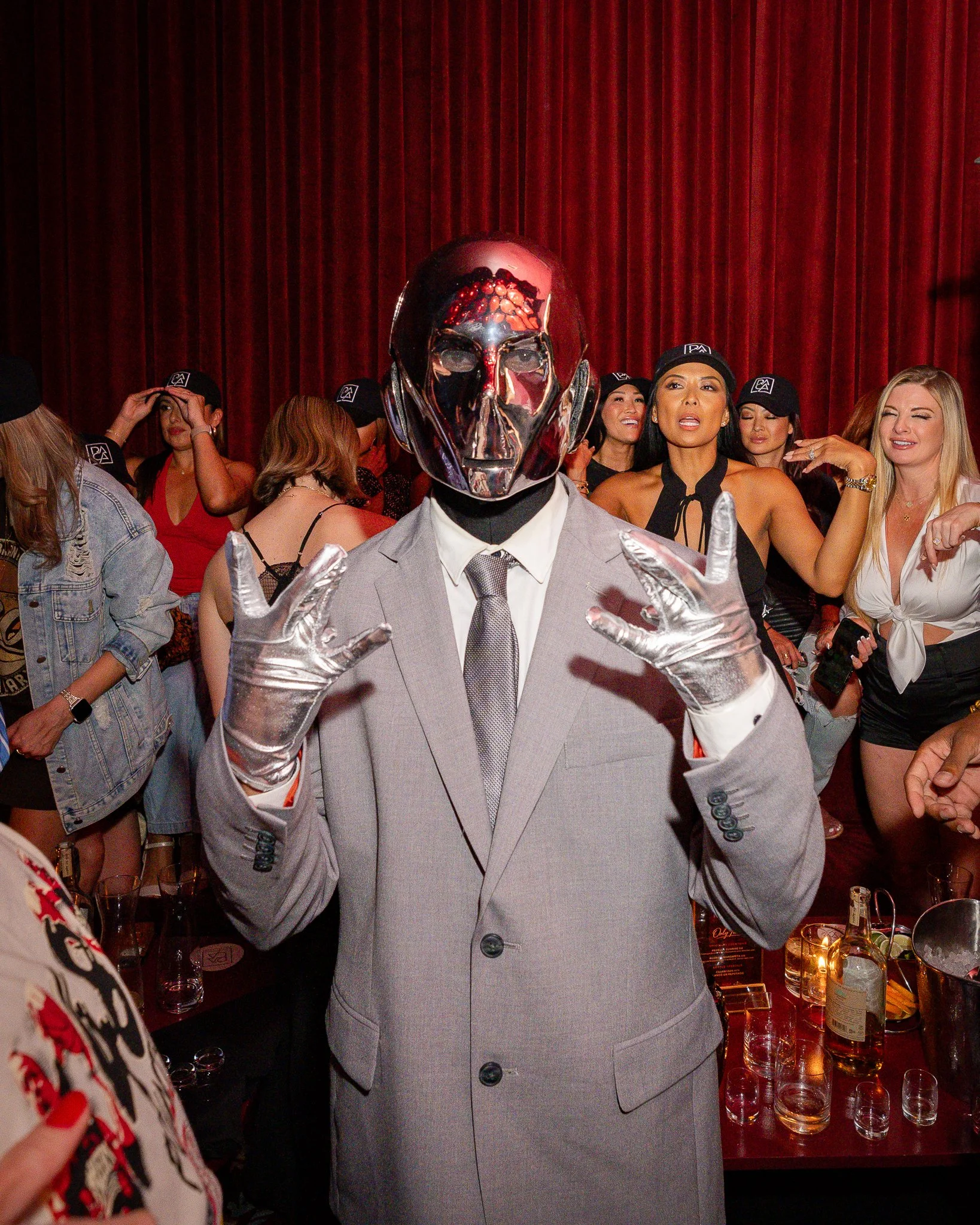 Person wearing a silver mask resembling a skull and a light-colored suit at a party, surrounded by women with drinks and illuminated by string lights in front of a red curtain.