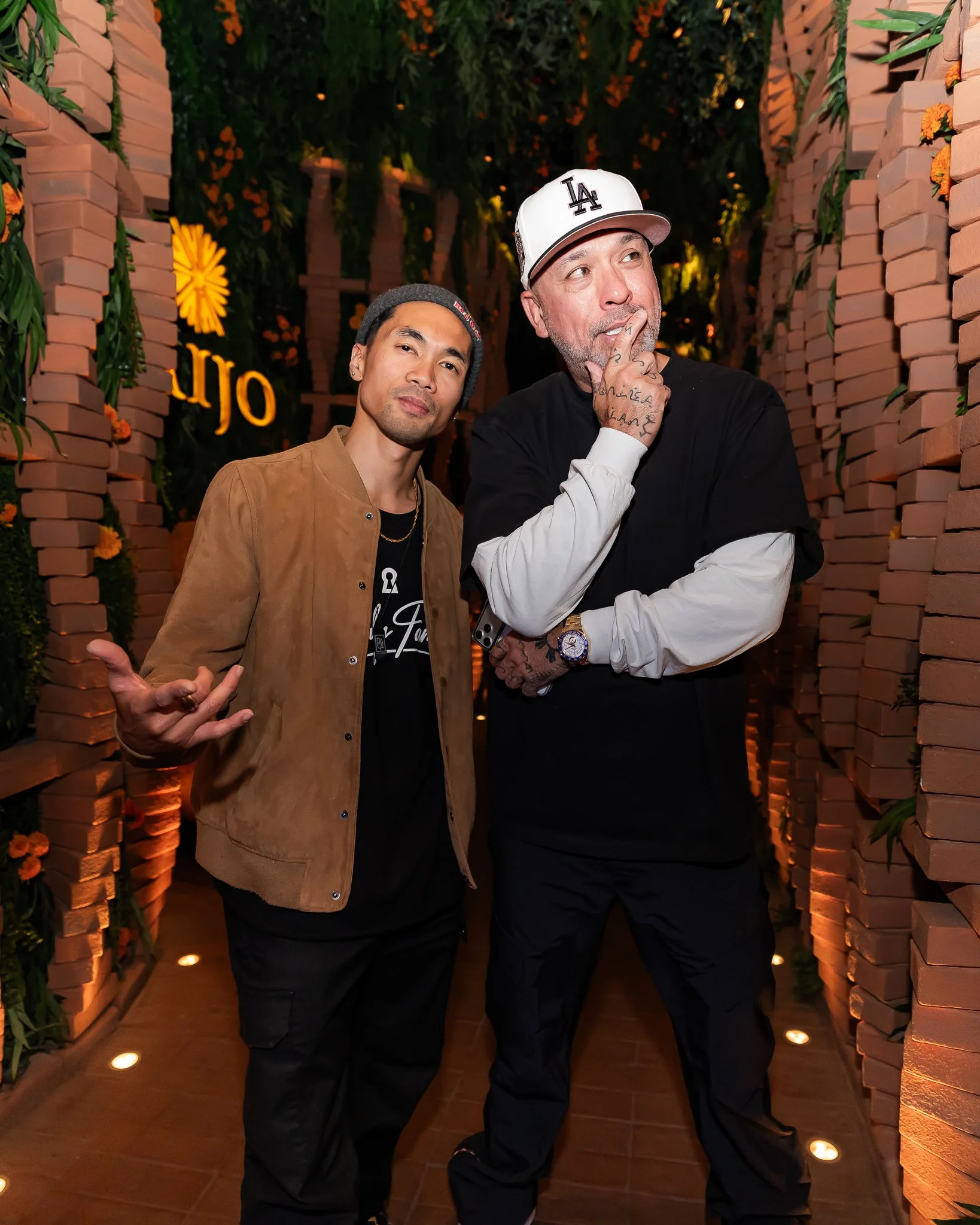 Two men posing in a decorative, brick-lined corridor with green plants and hanging flowers, illuminated by warm lighting. One man is making a shushing gesture, and the other is standing with a relaxed expression.