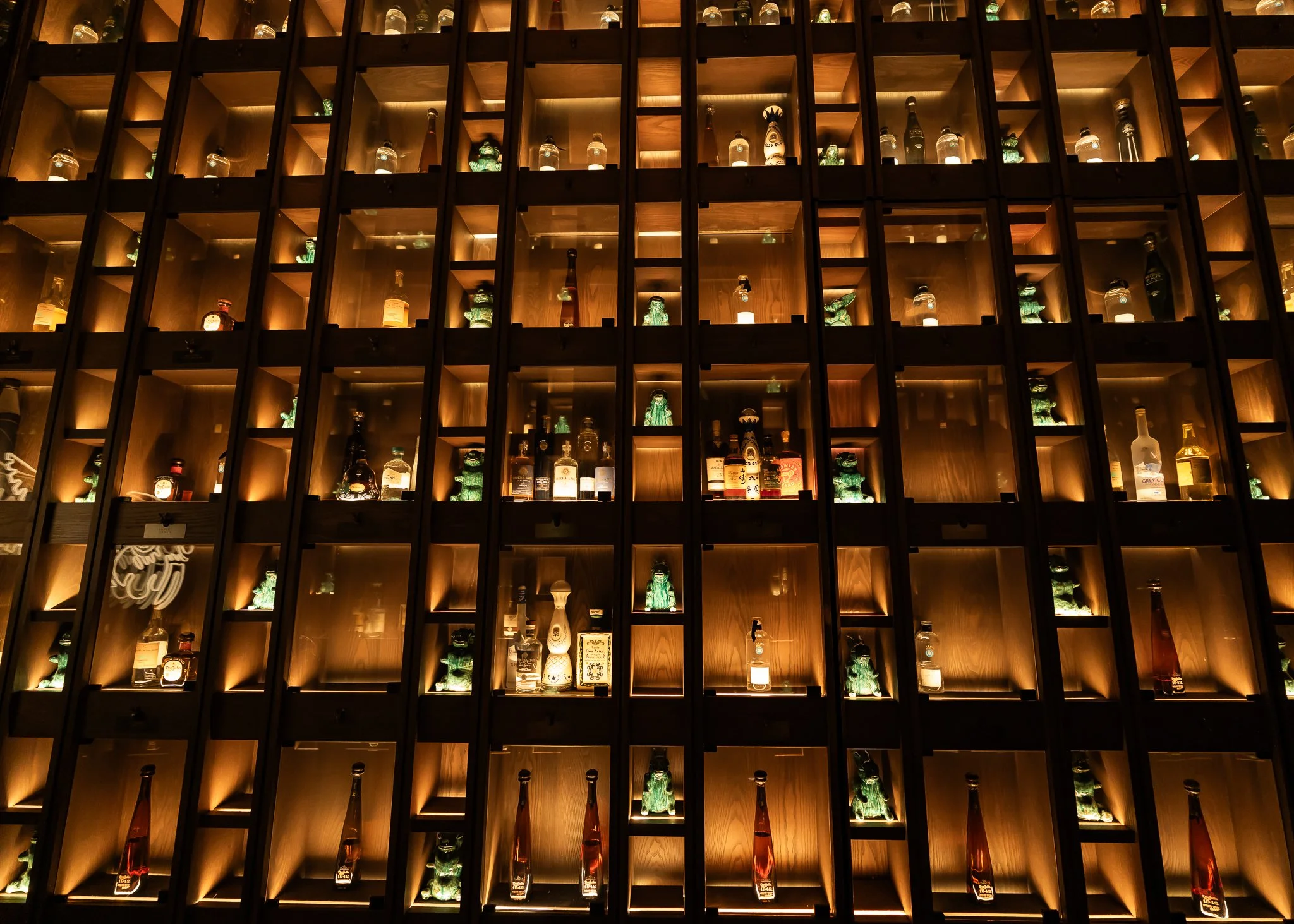View of a large wall with multiple compartments displaying various bottles of alcohol, illuminated with warm lighting.