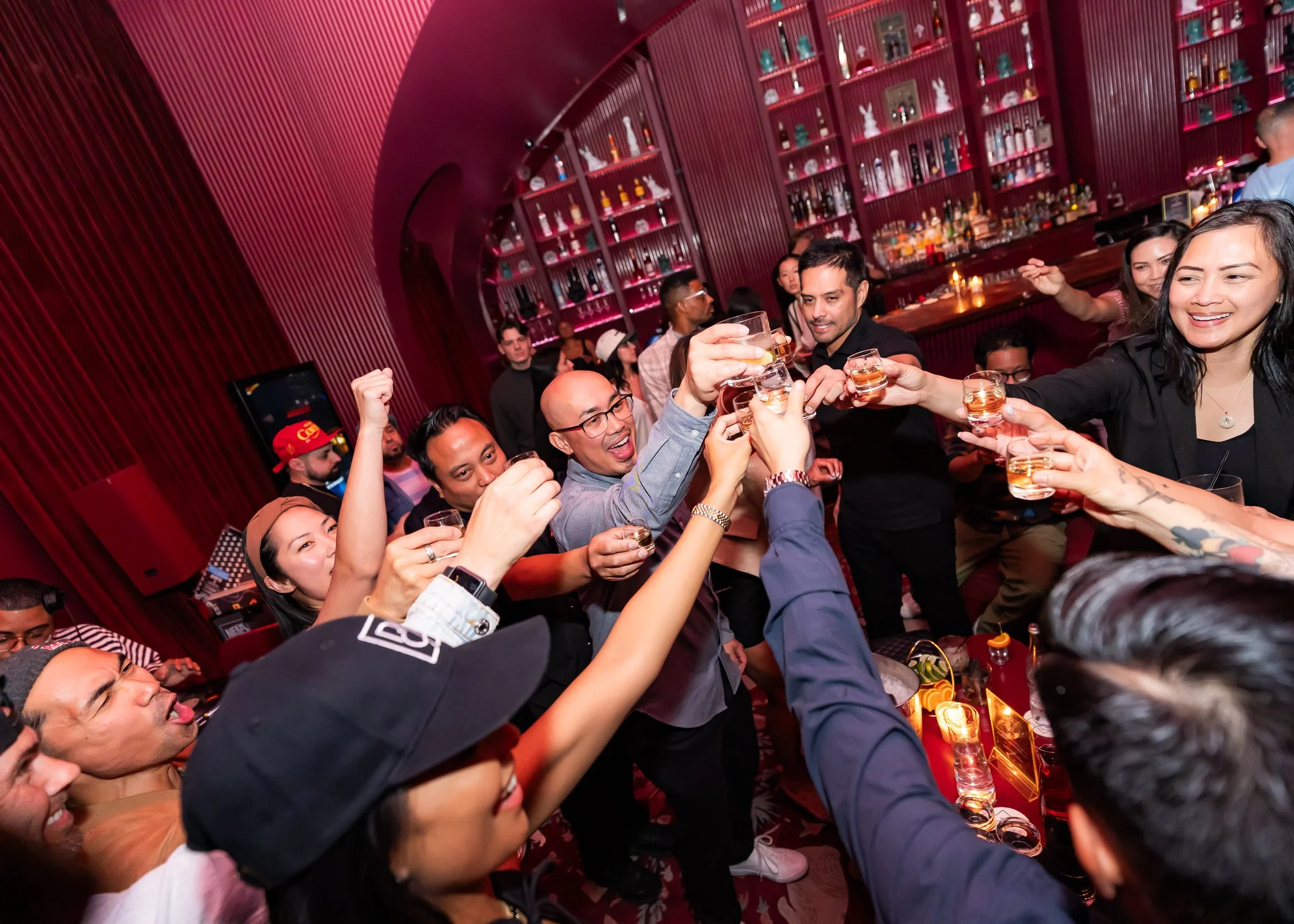 People at a bar celebrating, raising glasses, with a red interior and shelves full of bottles in the background.