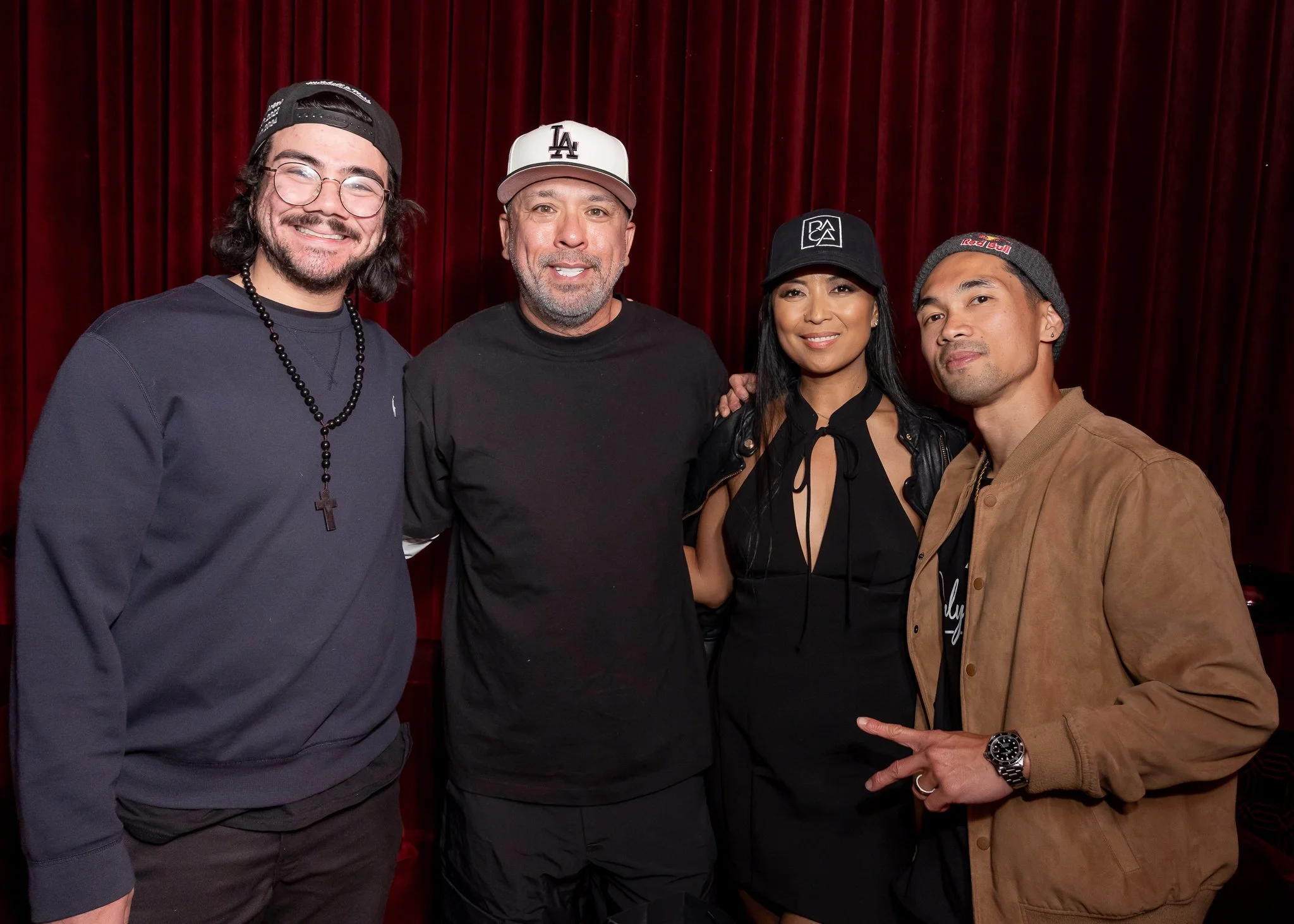 Four people standing close together in front of red curtains, smiling at the camera. The group includes three men and one woman, all casually dressed with hats or accessories.