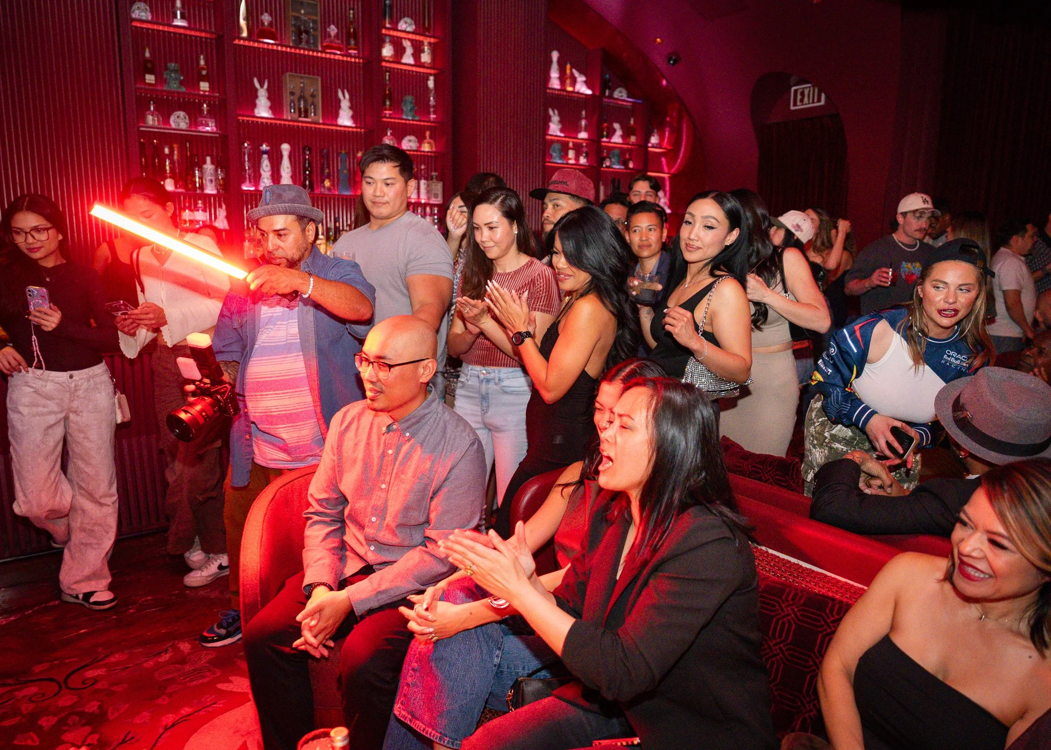 People gathered in a dimly lit bar or club with red lighting and shelves of bottles on the wall, some engaged with their phones, a man holds a red laser pointer, and others are clapping or watching something out of frame.