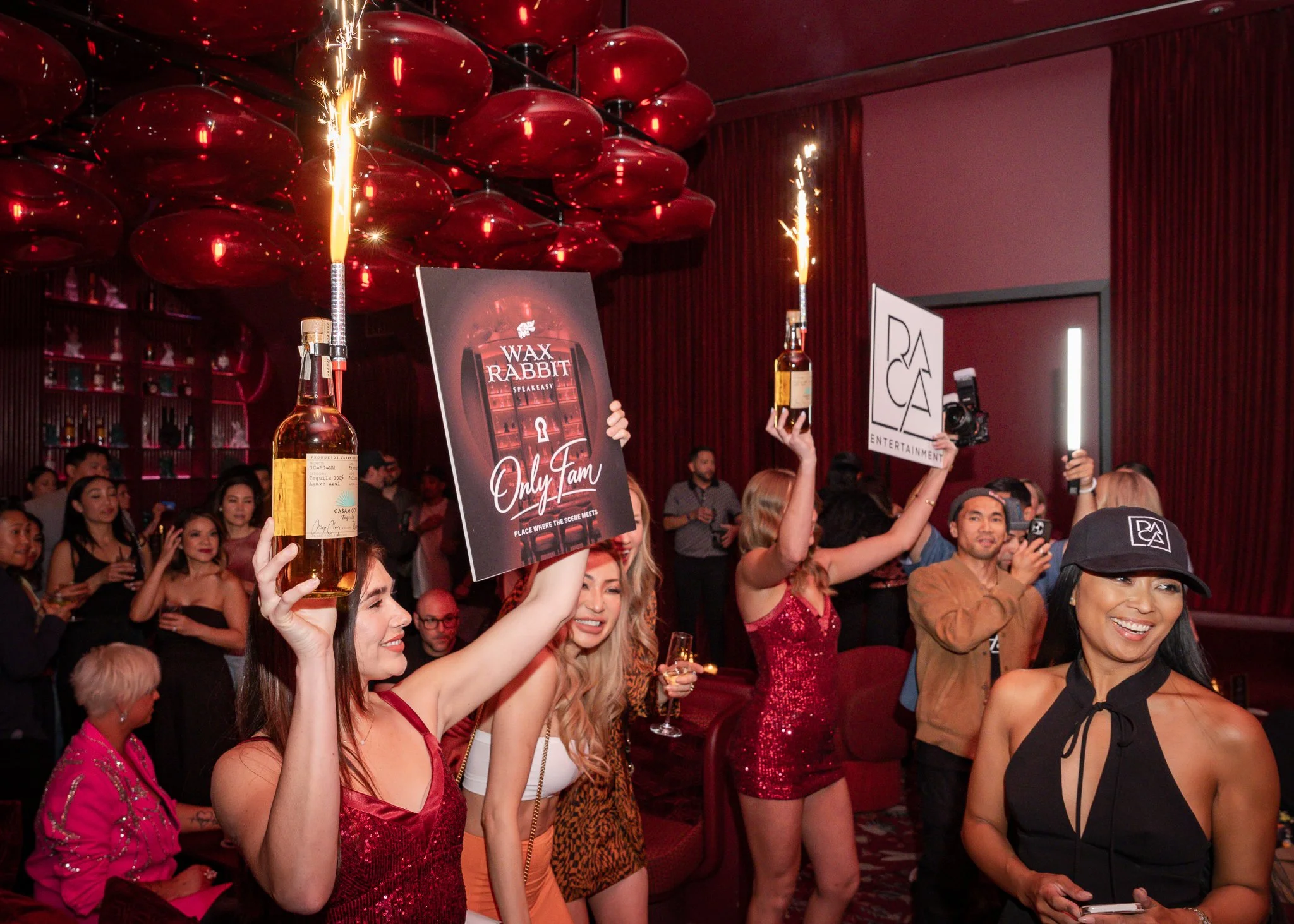 Party scene with people holding bottles and sparklers, decorated with red balloons and posters, in an indoor setting.