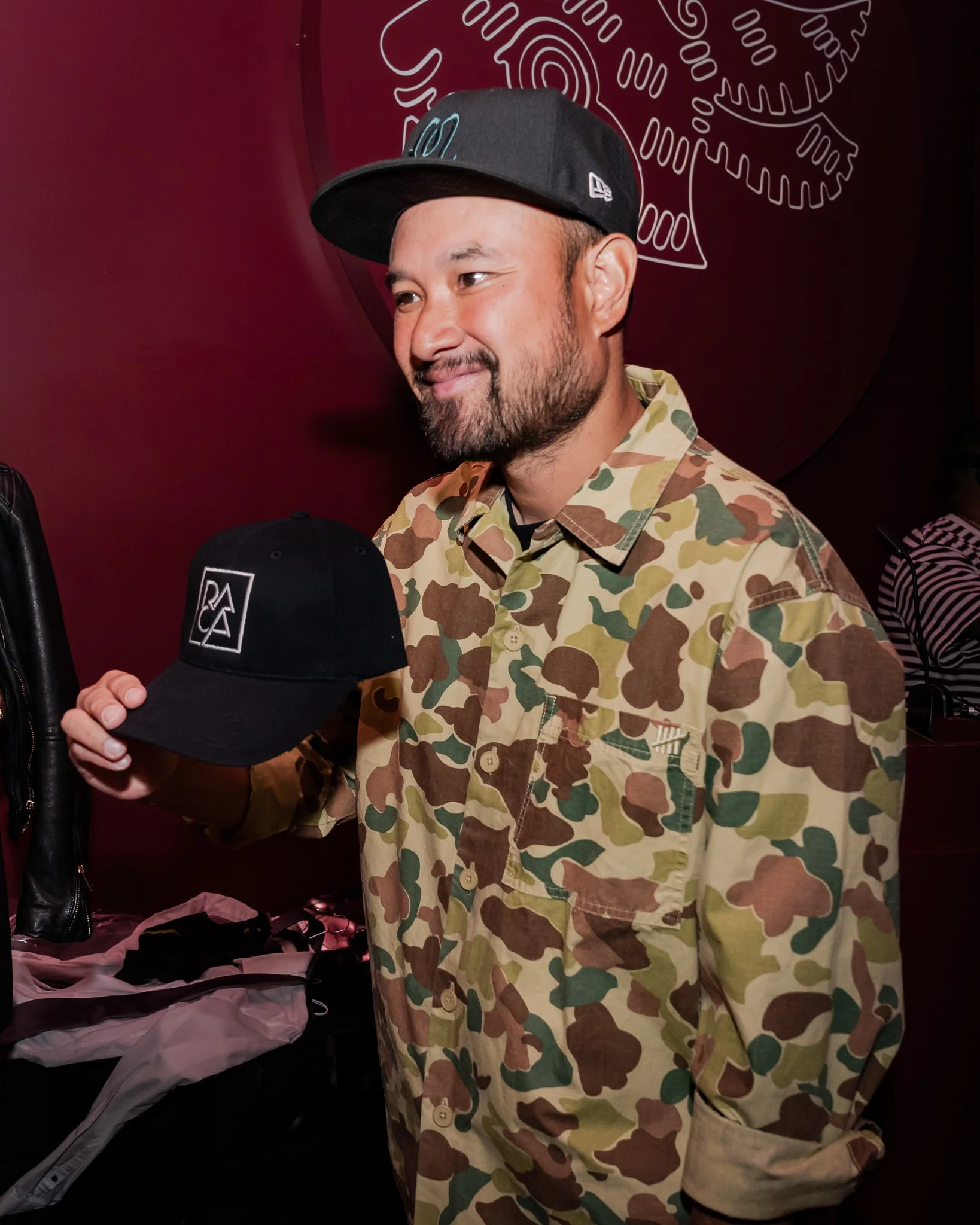 A man named Phi from Jabbawockeez with a beard wearing a black cap and camouflage shirt, smiling, standing in front of a maroon wall with a white line drawing of gears.
