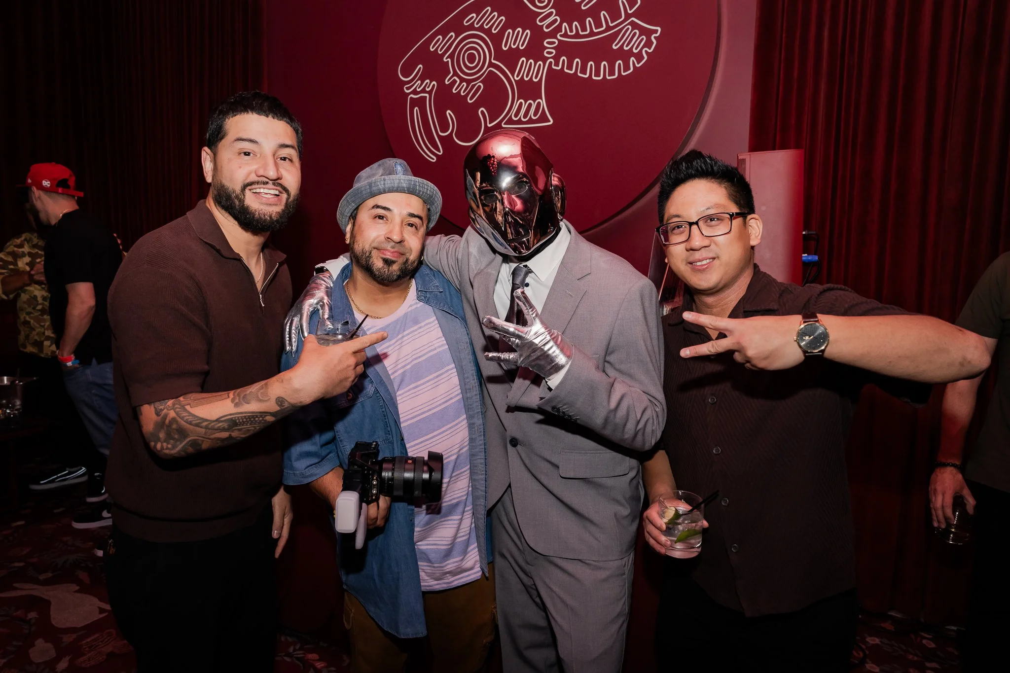 Four men posing together at a party, one wearing a silver mask and a suit, others dressed casually, all smiling and holding drinks.