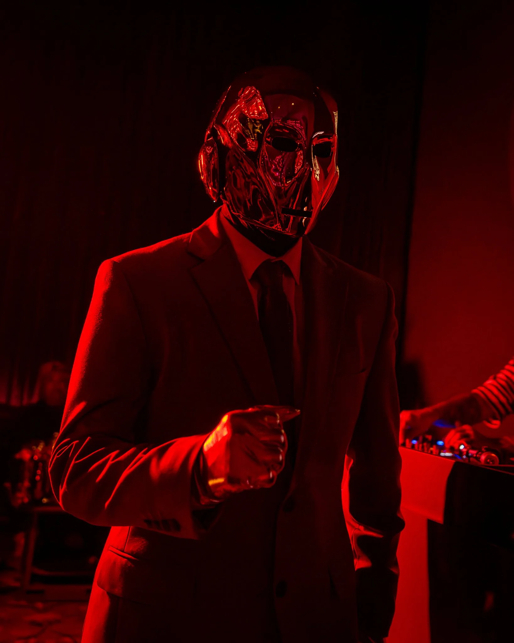 A person wearing a reflective metallic mask and a suit, illuminated by red lighting, standing in a dark room with DJ equipment visible in the background.