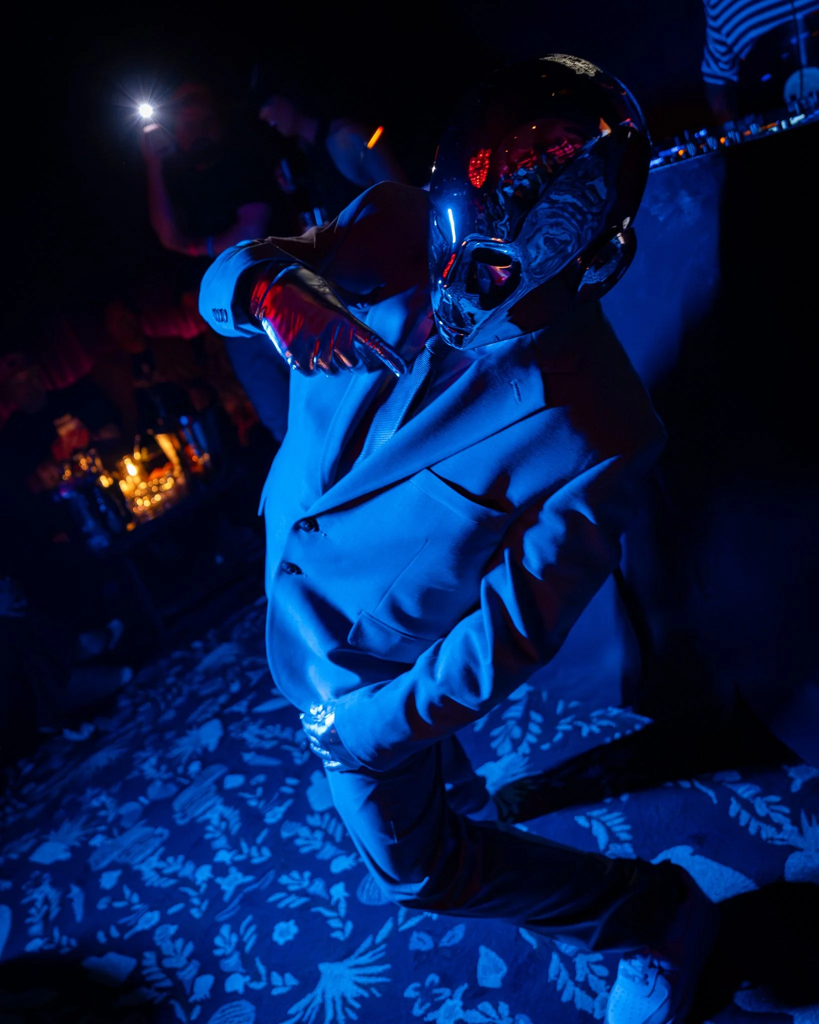 Person sitting in a dark room illuminated by blue and red lights, wearing gloves and a patterned shirt, with a glossy black helmet reflecting the surroundings, and other people visible in the background.