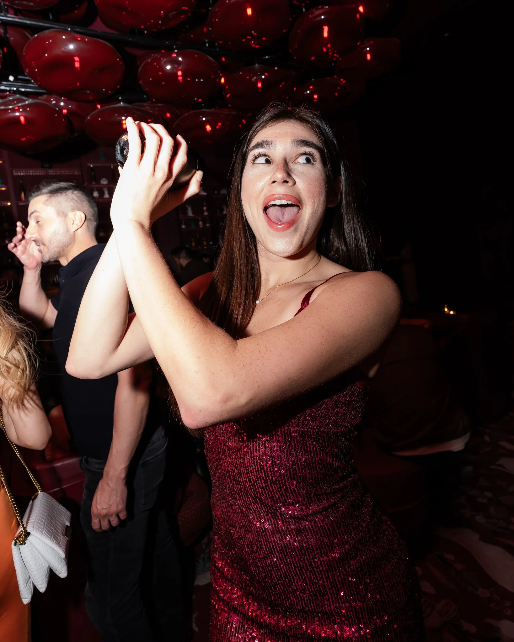 A young woman in a red sequin dress holding a glass at a celebration or party, smiling and looking to the side. A man is in the background, facing away, with his eyes closed and hand near his face.