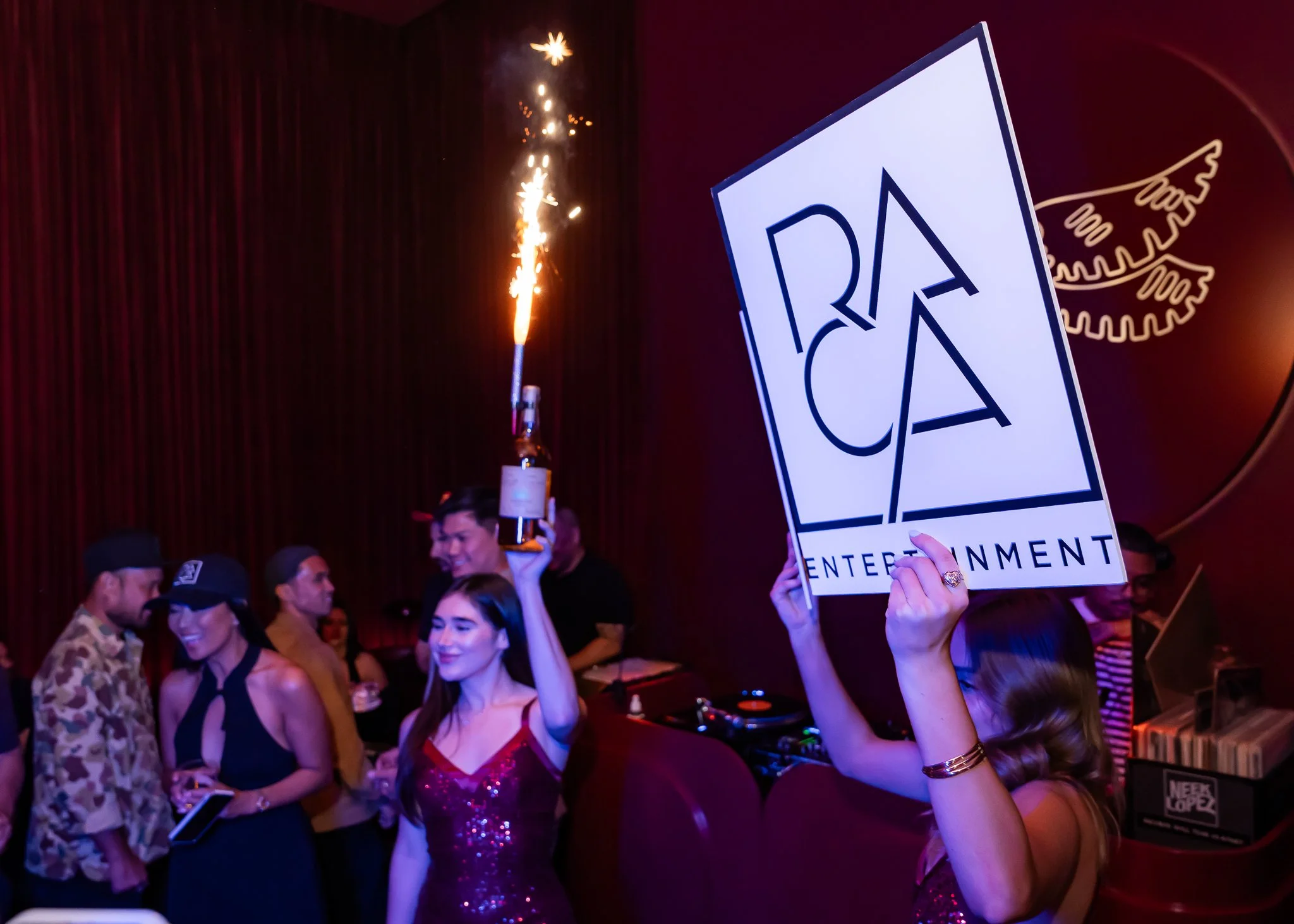 People celebrating at an event, holding a sign that says 'RAA ENTERTAINMENT,' with a sparkler in the background and a DJ setup visible.
