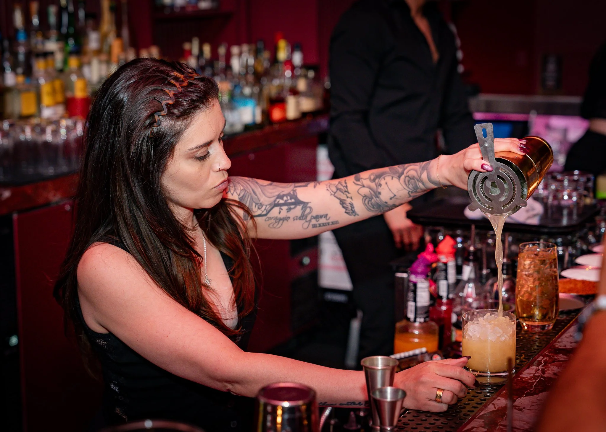 A woman with long dark hair and tattoos on her arm pours a drink with a gold-colored shaker at a bar counter, with various bottles on shelves in the background.
