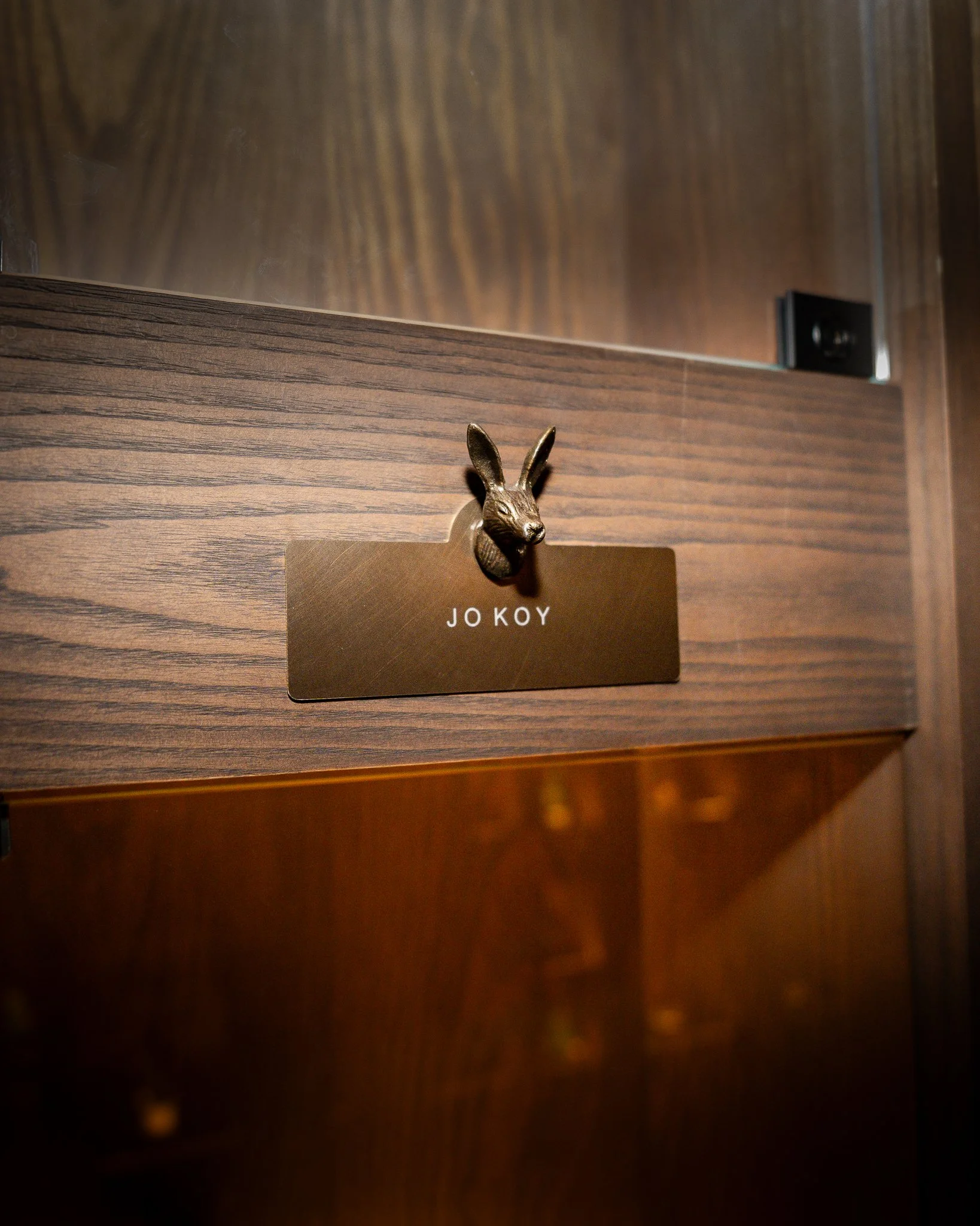 A dark wooden piece of furniture, possibly a cabinet or drawer, with a decorative rabbit figurine on top, which is holding or attached to a gold card with the text 'JOKOY' on it.