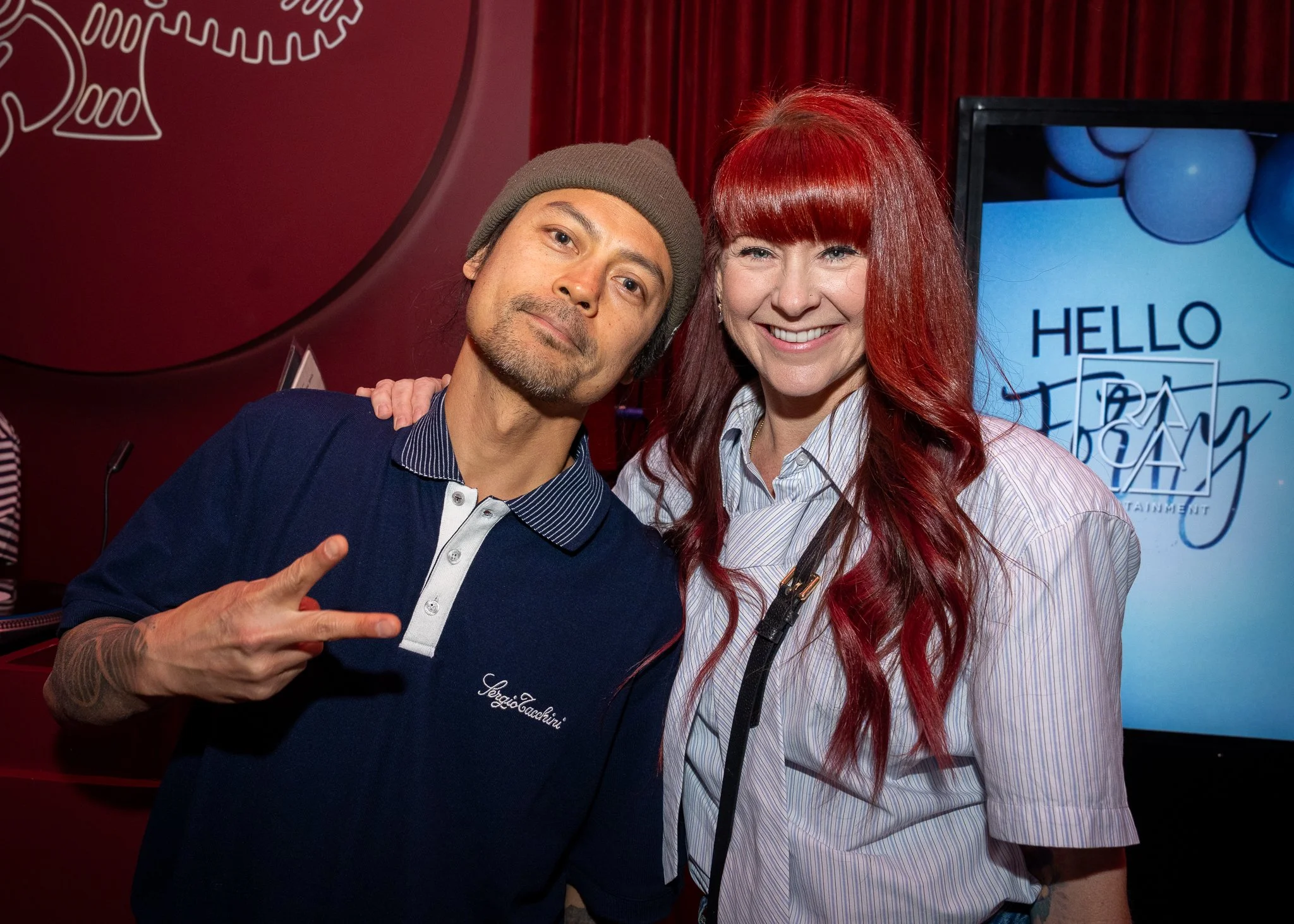 Two people, a man and a woman, smiling and posing for a photo indoors. The man wears a grey beanie and a dark blue shirt, and the woman has long red hair and is wearing a striped shirt. They are standing in front of a red curtain with a television sc