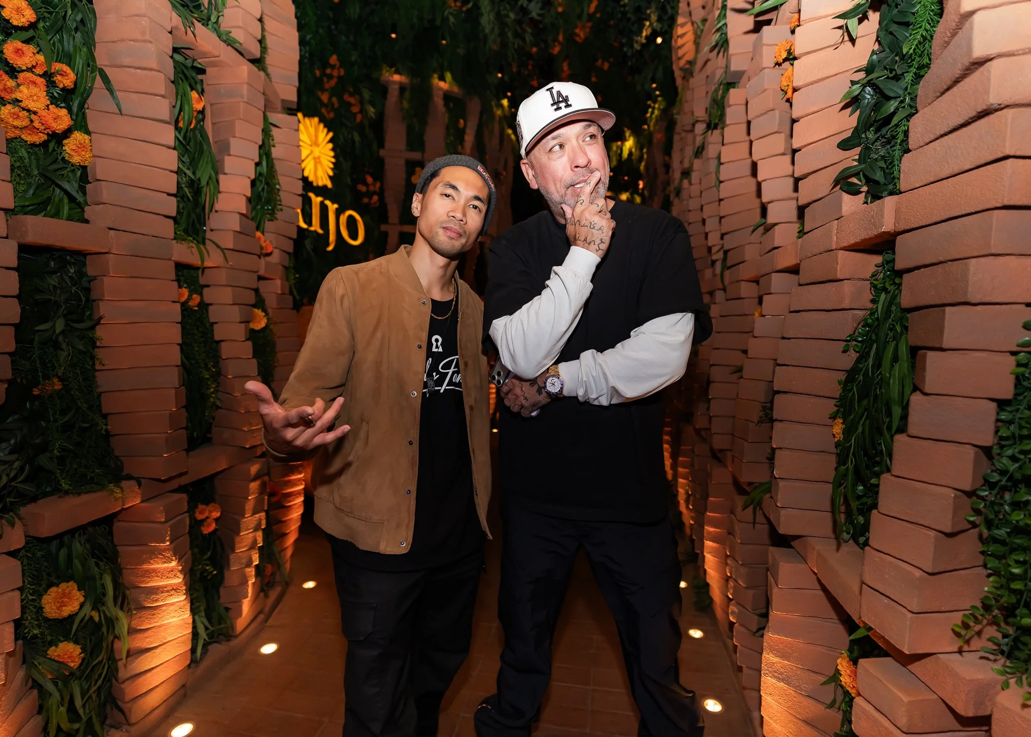 Two men posing in a decorated brick corridor with flowers and warm lighting, one wearing a white LA baseball cap and the other in a brown jacket and black beanie.