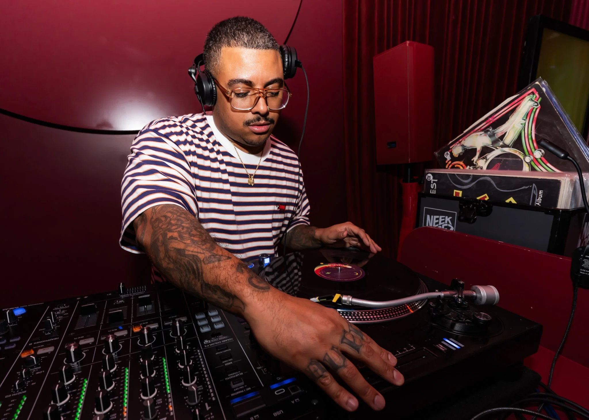 A DJ with short hair, glasses, and tattoos wearing a striped shirt, is mixing music with DJ equipment in a red-lit room.