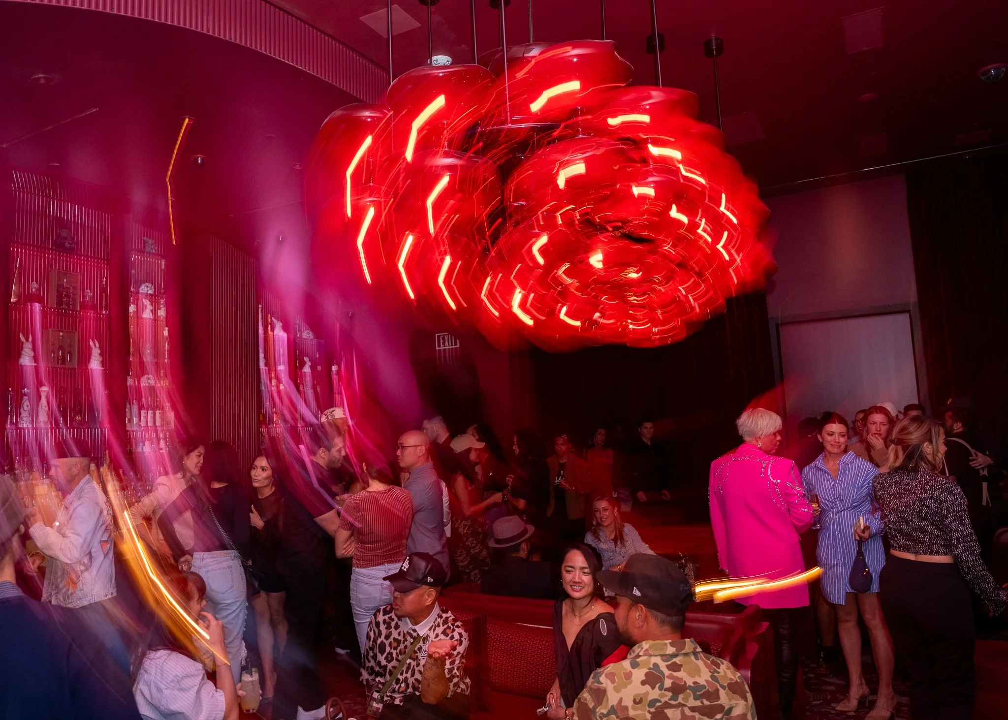A crowded indoor event with people socializing and a large red modern chandelier overhead, with some blurred motion effects.