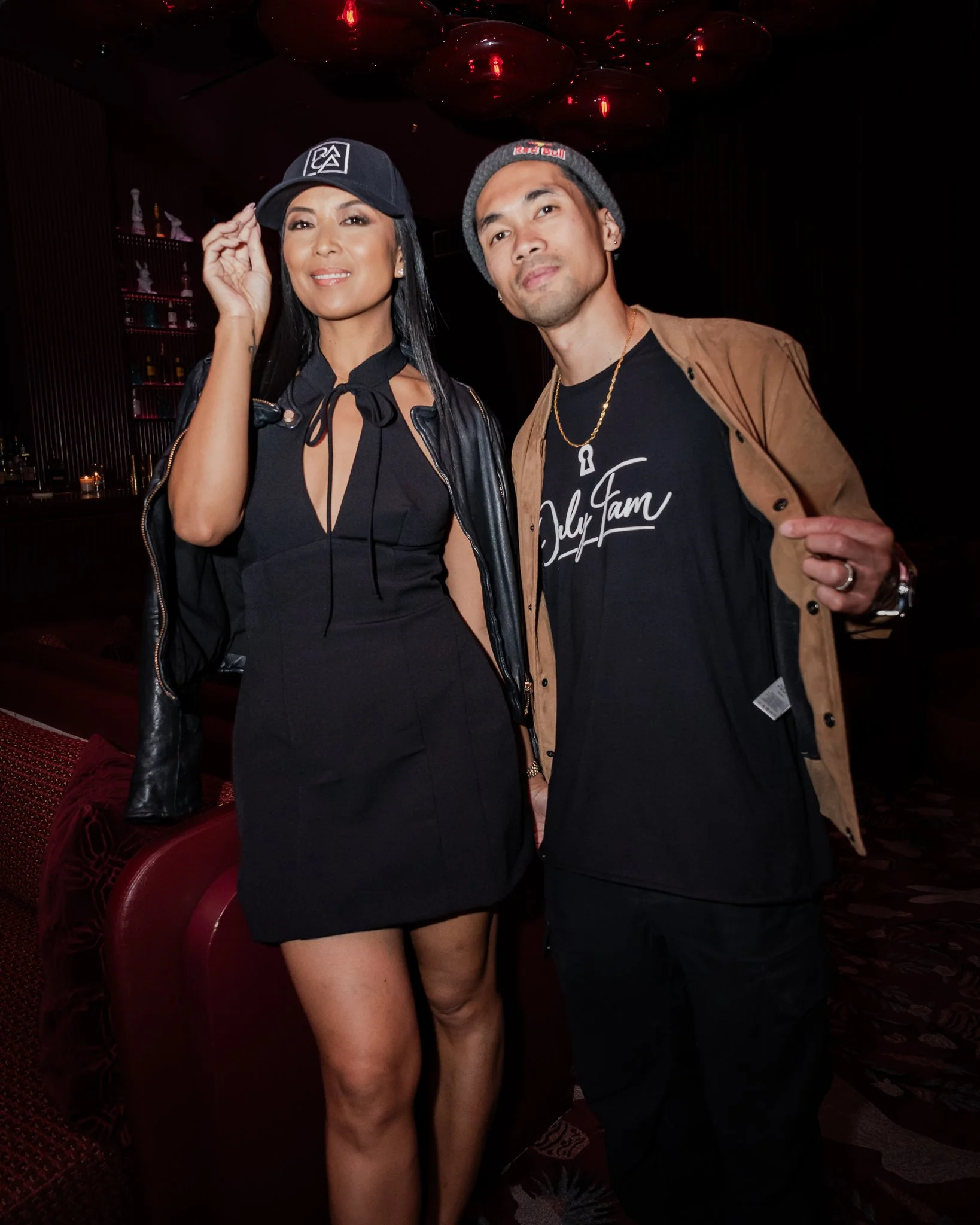 A woman in a black dress with a thigh-high slit and a man in a brown jacket poses together in a dimly-lit setting with red decor elements and shelves of bottles in the background.