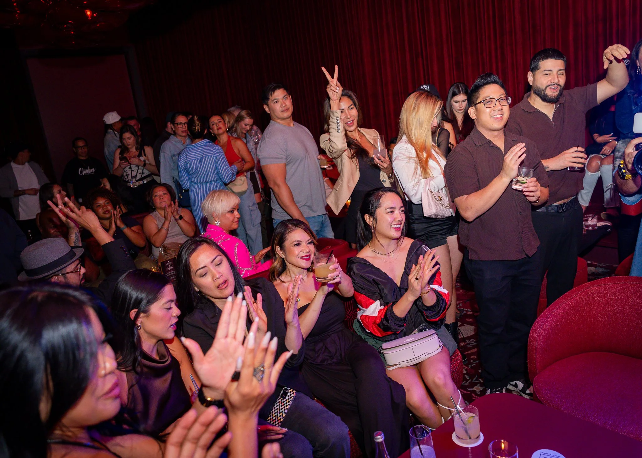 A group of people at a party, some standing and some seated, with drinks in hand, in a dimly lit room with red curtains.