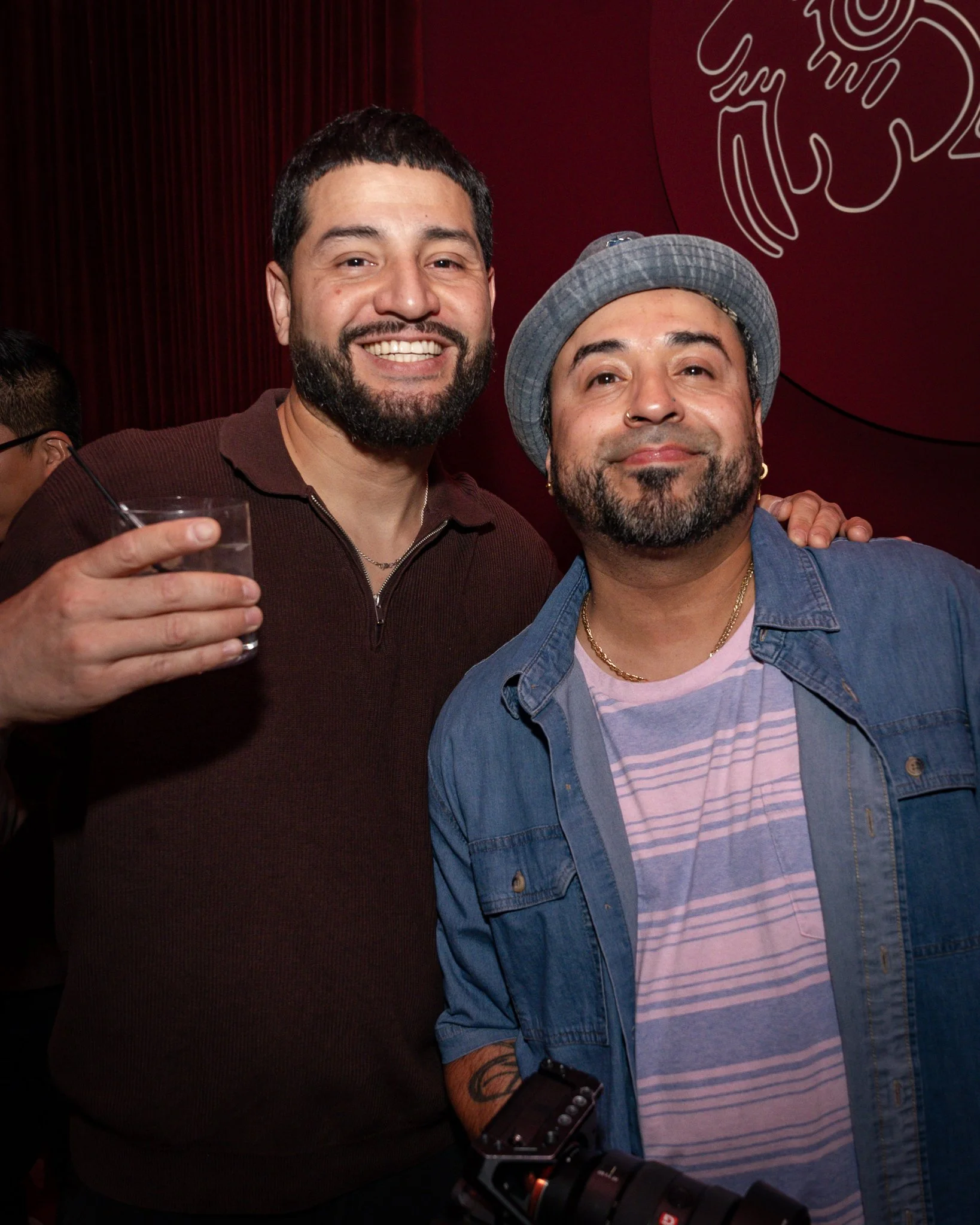 Two men smiling for the camera at a social event, with one holding a drink and the other with his arm around his friend, against a dark red background with a line art graphic.