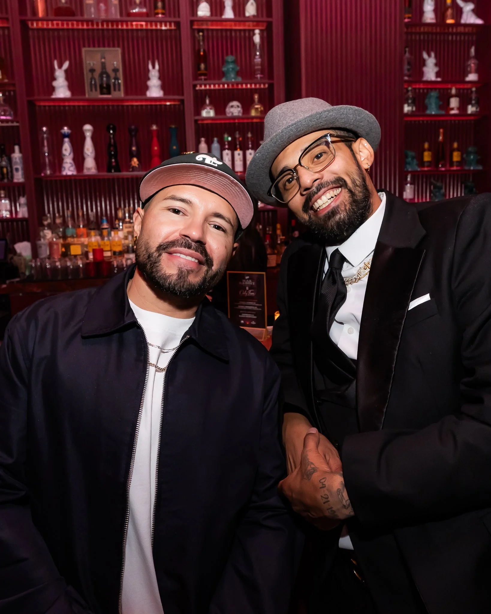 Two men smiling and shaking hands in a bar or lounge with shelves of bottles and decorative items in the background.