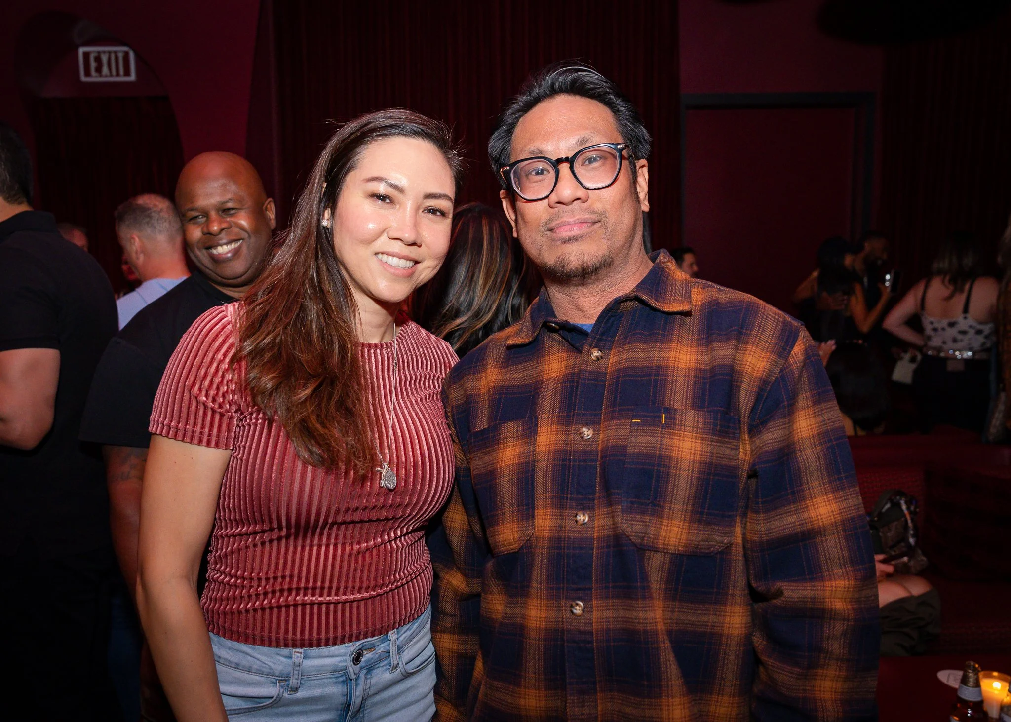 A group of people at an indoor event, with two people in the foreground, smiling and looking at the camera. The woman on the left has long, wavy hair, wearing a red, striped top and light blue jeans. The man on the right has black hair, glasses, and 
