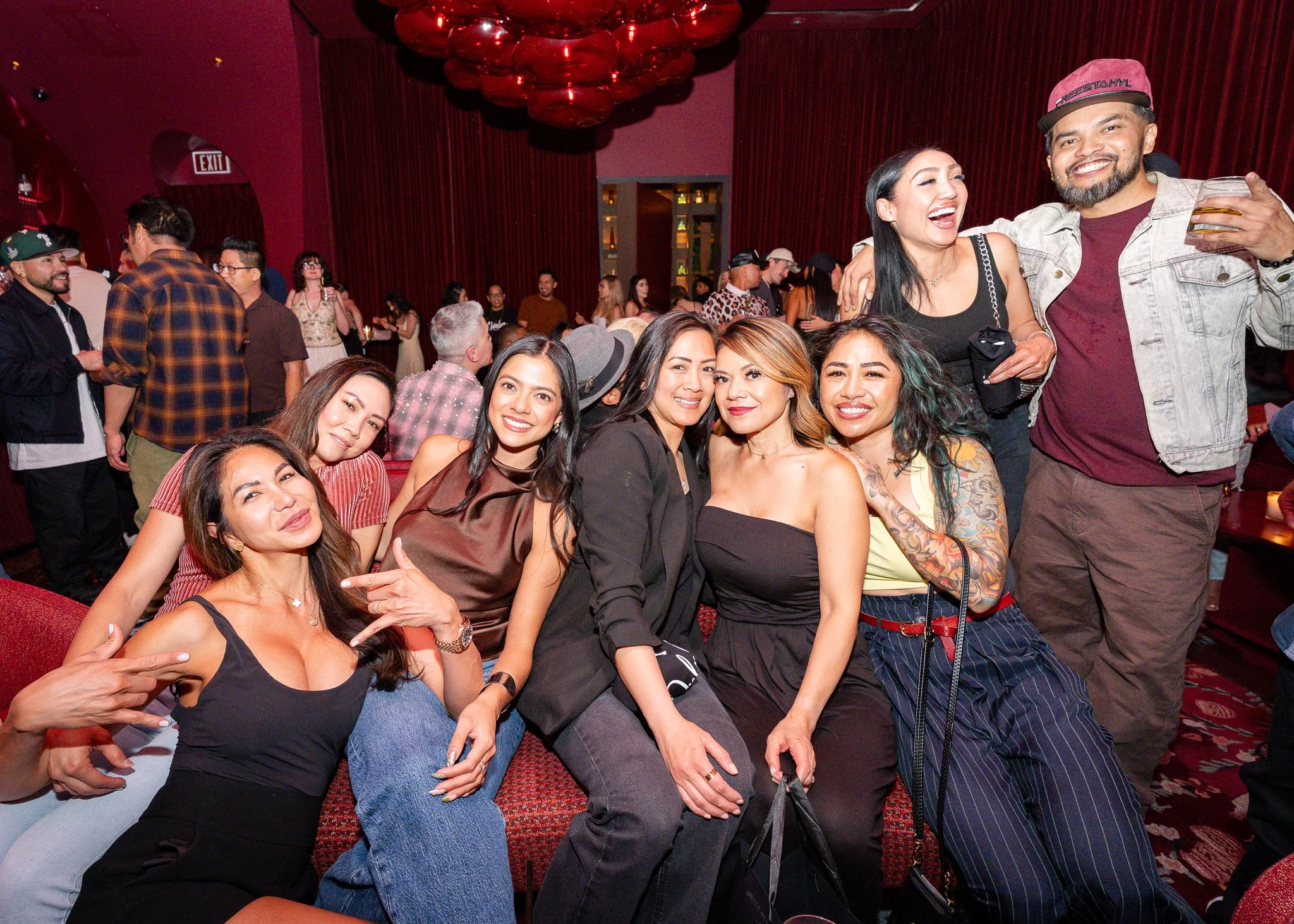 Group of people enjoying a social event in a dimly lit venue with red interior decor, some seated on red bench seats and others standing and chatting, smiling, and holding drinks.
