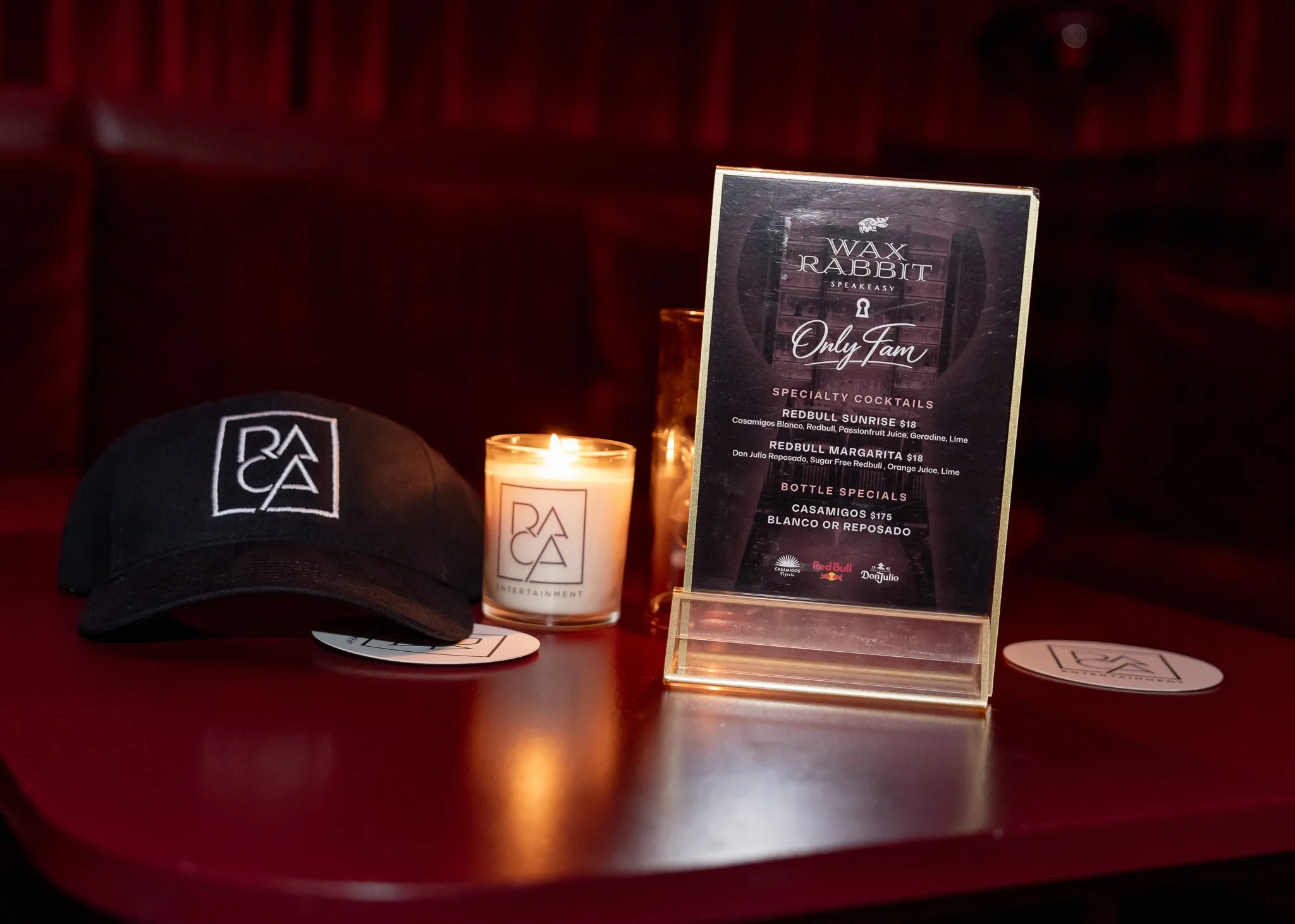 A display table with a black cap, a lit candle, and a clear acrylic stand with a menu for Wax Rabbit Speakeasy cocktails, including Redbull Sunrise and Redbull Margarita, in a dimly lit setting with a red background.