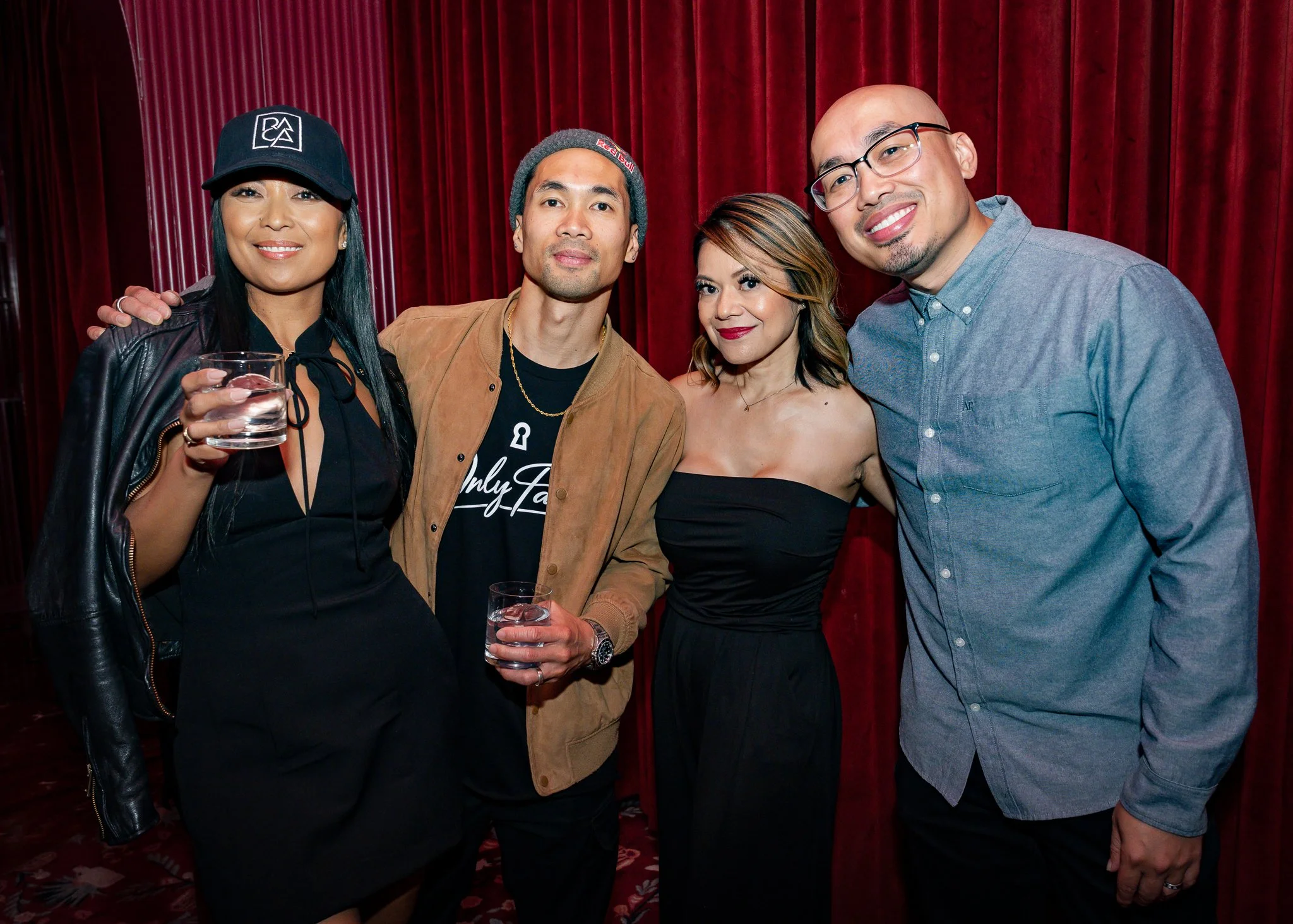 Group of five diverse people standing together in front of a red curtain, smiling, with one person holding a glass of water.