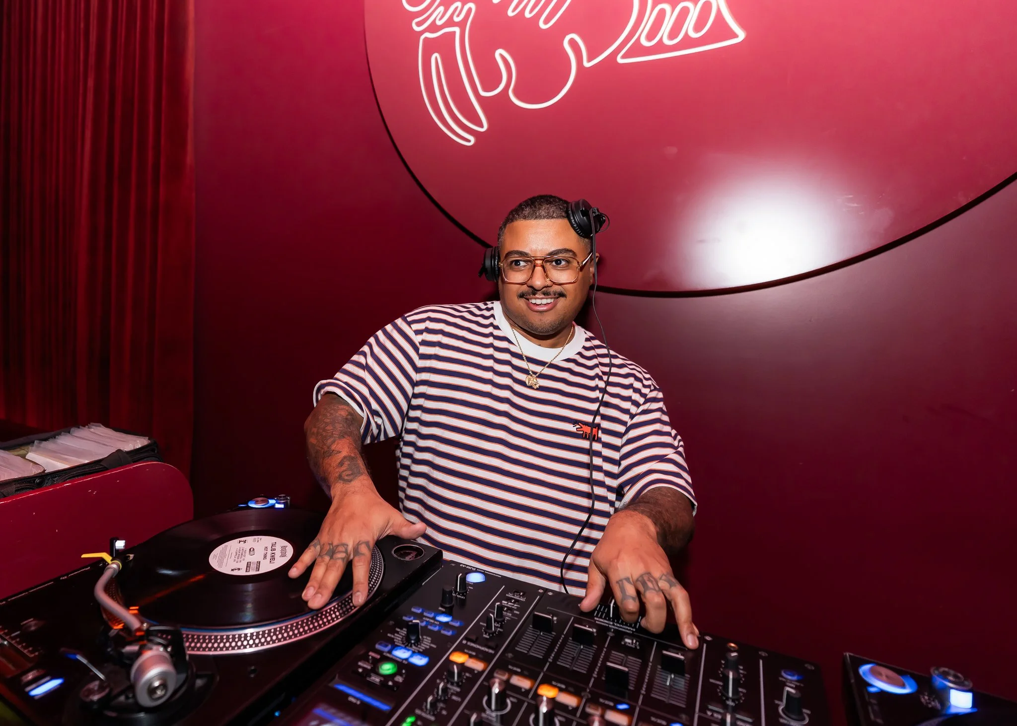 A man with tattoos on his arms, wearing glasses and a striped shirt, is DJing with a turntable and mixer at a red-themed venue. He's smiling and wearing headphones, with a neon sign behind him on a red wall.