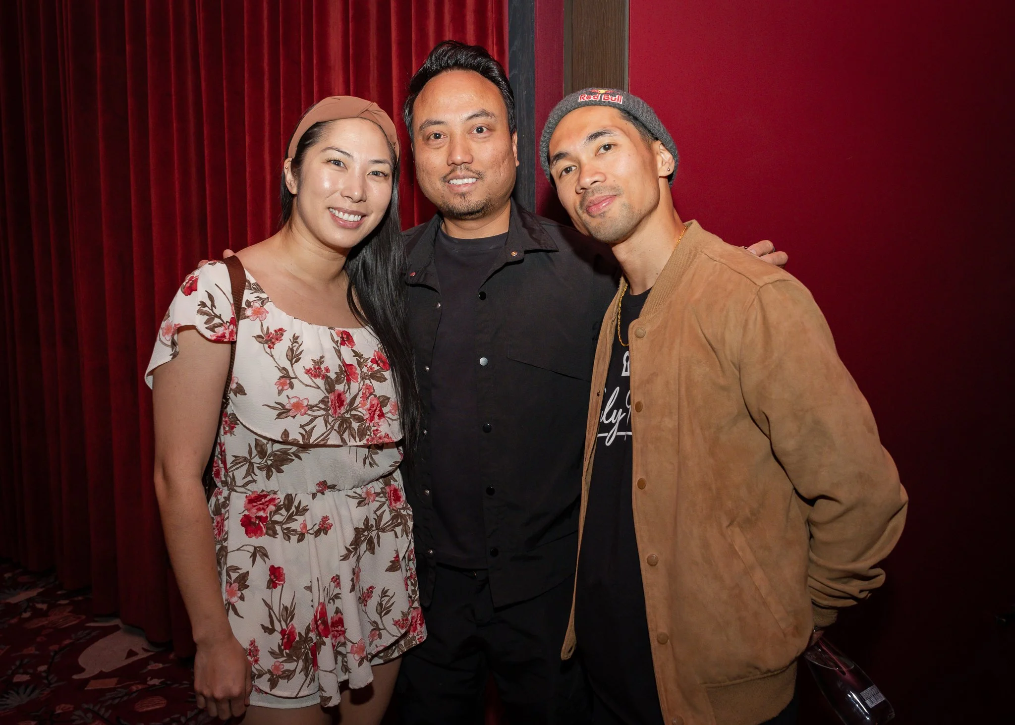 Three people standing close together, smiling, in front of a red curtain. The woman on the left is wearing a beige dress with a red and brown floral pattern and has long dark hair. The man in the middle has short black hair, facial hair, and is weari
